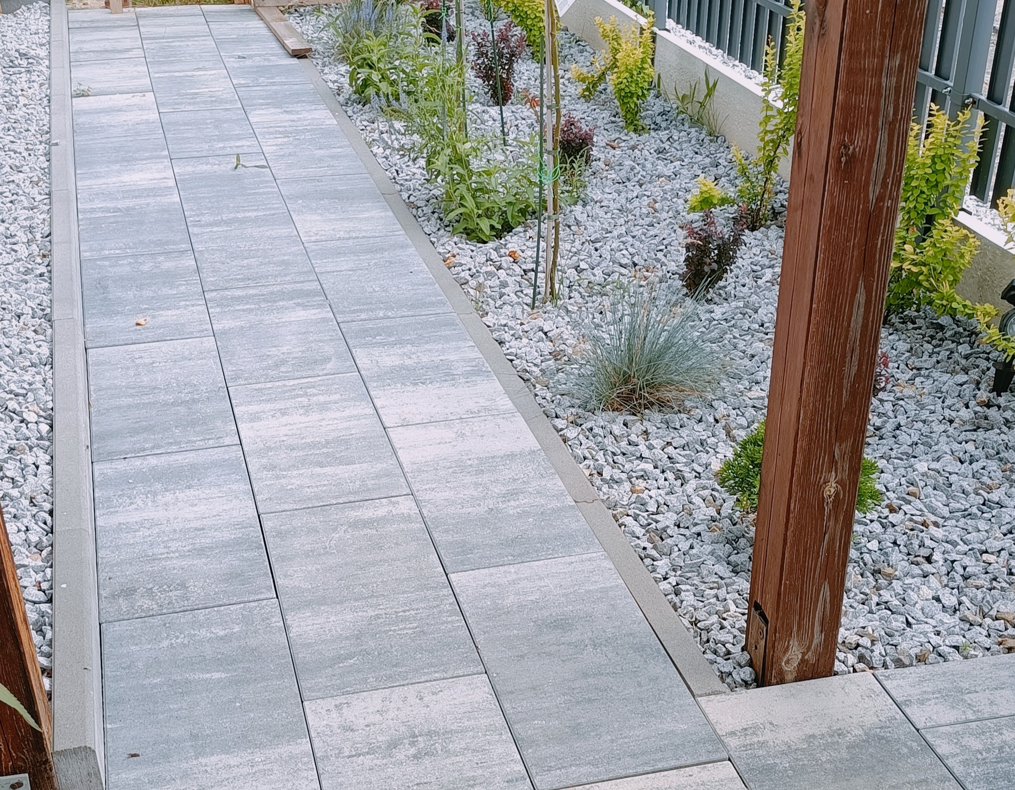 Ułożona ścieżka z szarych płyt betonowych, obok rabata z roślinami i kamieniami. Widoczny drewniany słupek i fragment metalowego ogrodzenia.