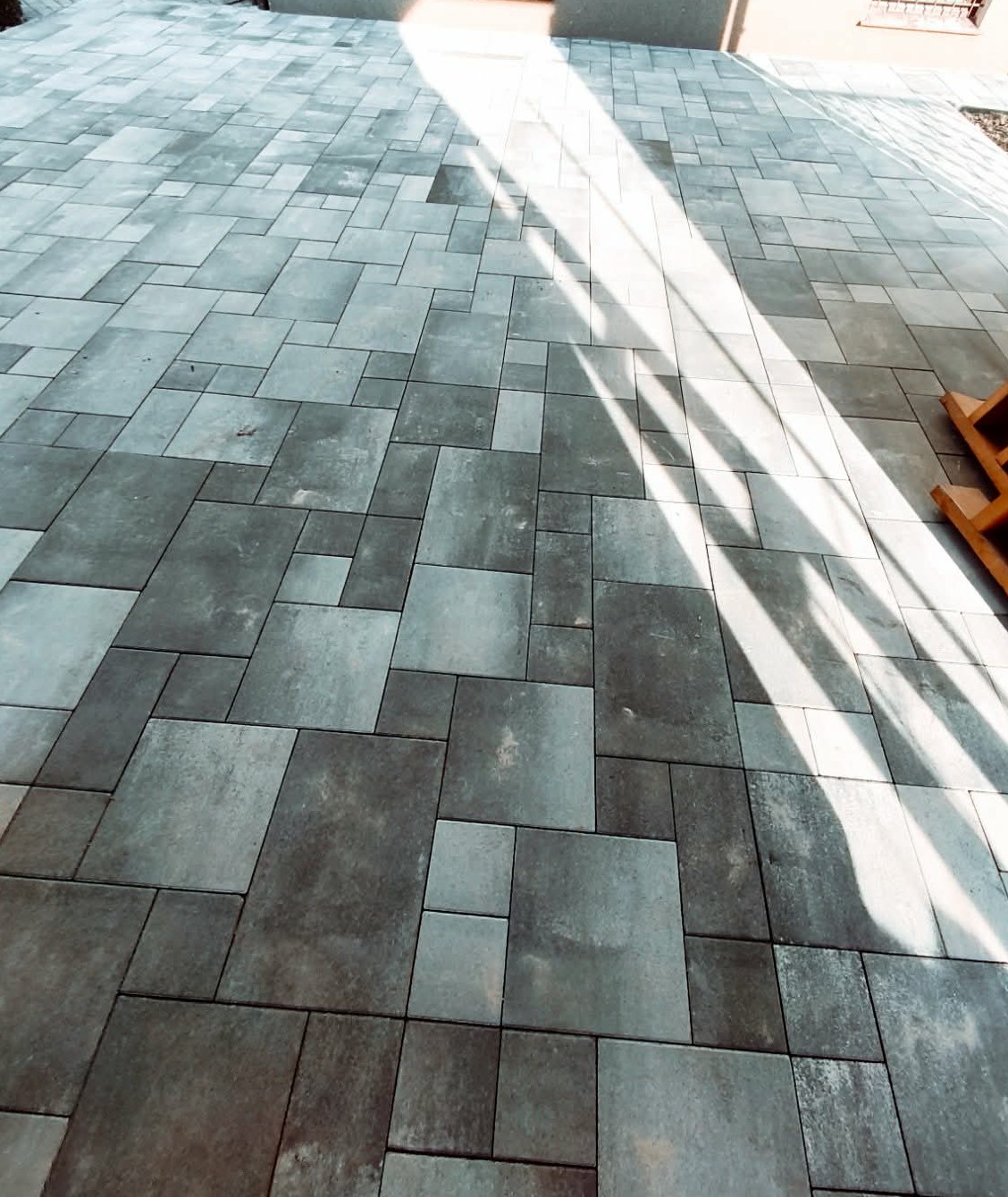 Ułożona kostka brukowa w odcieniach szarości, z wyraźnym wzorem i padającym światłem słonecznym. Widoczny fragment budynku w tle.