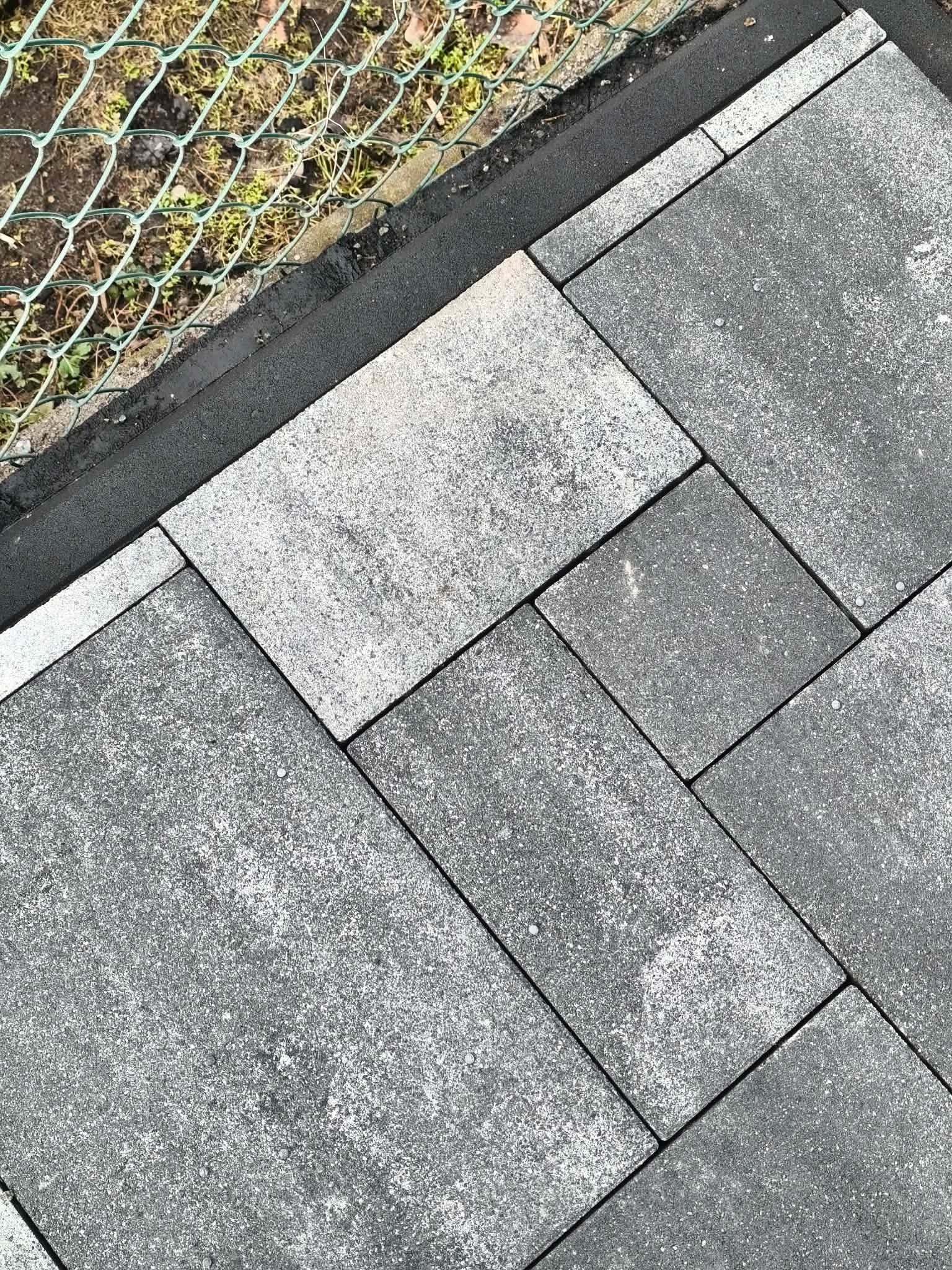 Ułożona, szara kostka brukowa w Radomierowicach, widoczna z góry, z fragmentem siatki ogrodzeniowej w tle. Kostka ułożona w regularny wzór, z widocznymi fugami.