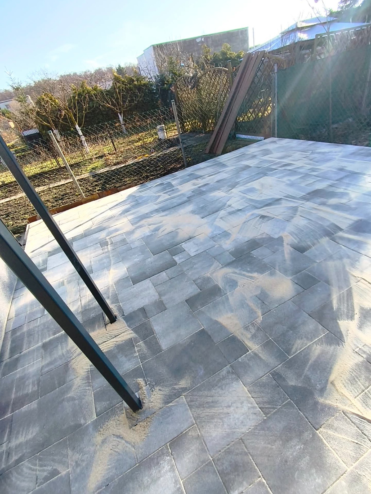 Ułożona, szara kostka brukowa na posesji, widok z góry. Elementy konstrukcyjne w lewym dolnym rogu. W tle siatka ogrodzeniowa i zieleń. Słoneczny dzień.