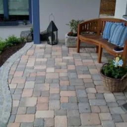 Ułożona kostka brukowa w odcieniach szarości i beżu tworzy estetyczny taras z ławką, latarnią i roślinami. Obrzeża z kostki granitowej.