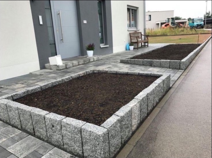 Granitowe obrzeża rabat i ścieżka z kostki przy domu. W tle widoczny budynek i sprzęt budowlany. Obrzeża wykonane z szarej kostki granitowej.