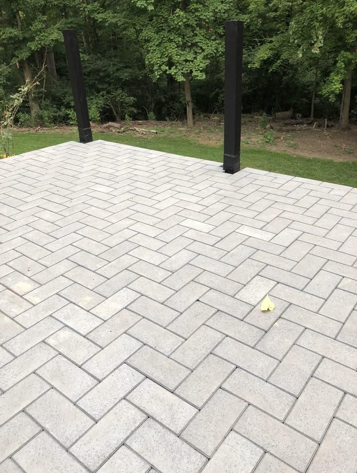 Ułożona szara kostka brukowa w jodełkę, z dwoma czarnymi słupkami w tle i zielenią. Widoczny pojedynczy, żółty liść na powierzchni kostki.