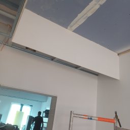 NK ProBud Usługi Ogólnobudowlane Nikodem Kostuch - Montaż sufitu podwieszanego z płyty gipsowej, widoczne profile metalowe, szpachlowane łączenia. W tle pracownik i rusztowanie.