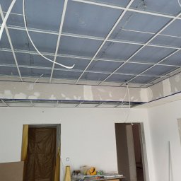 NK ProBud Usługi Ogólnobudowlane Nikodem Kostuch - Montaż sufitu podwieszanego z płyt gipsowo-kartonowych na metalowej konstrukcji. Widoczne przewody i częściowo wykończone ściany w jasnym pomieszczeniu.