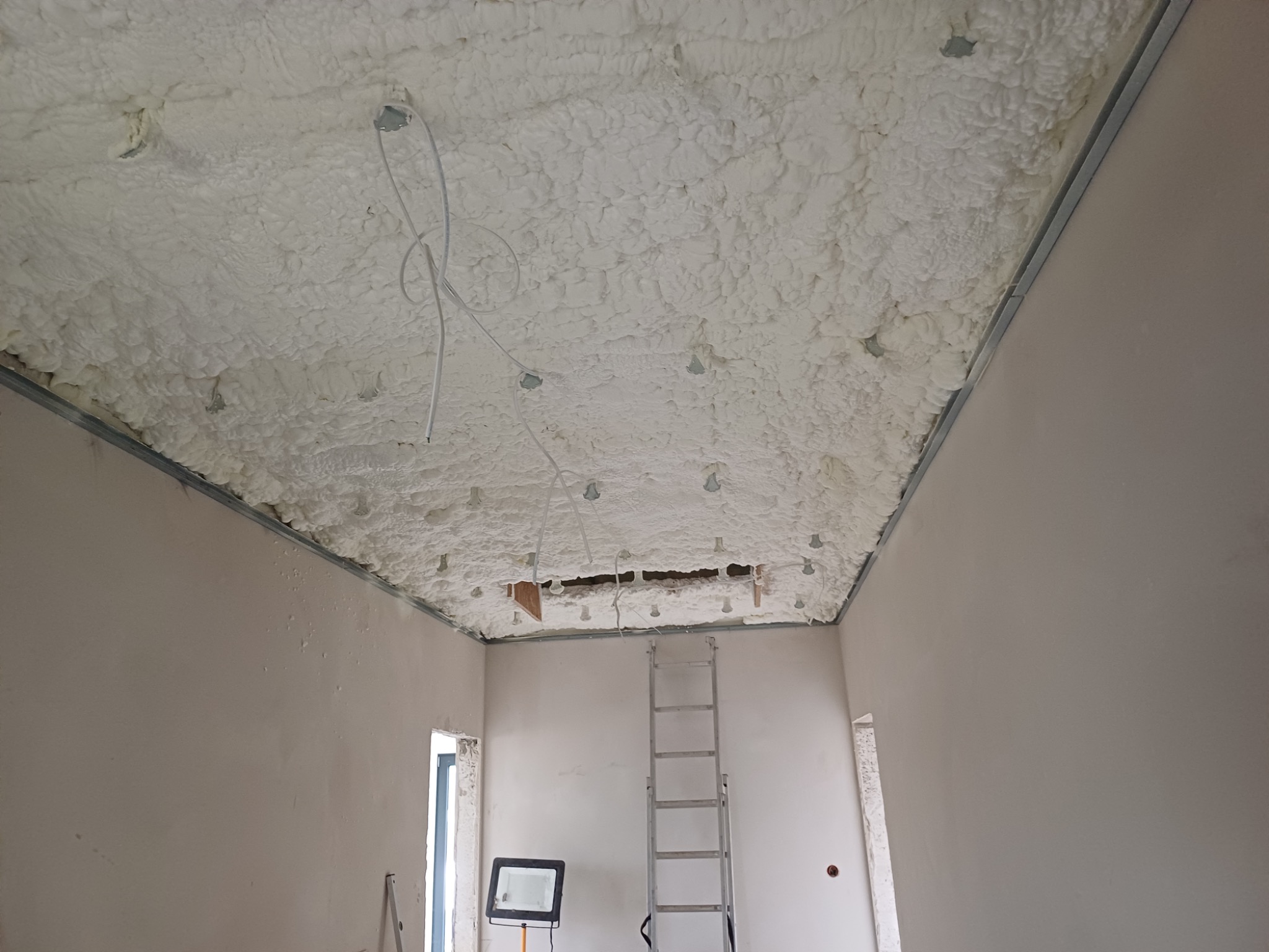 Piana izolacyjna na suficie z widocznymi przewodami i metalową konstrukcją. W tle drabina i oświetlenie halogenowe. Etap prac wykończeniowych.