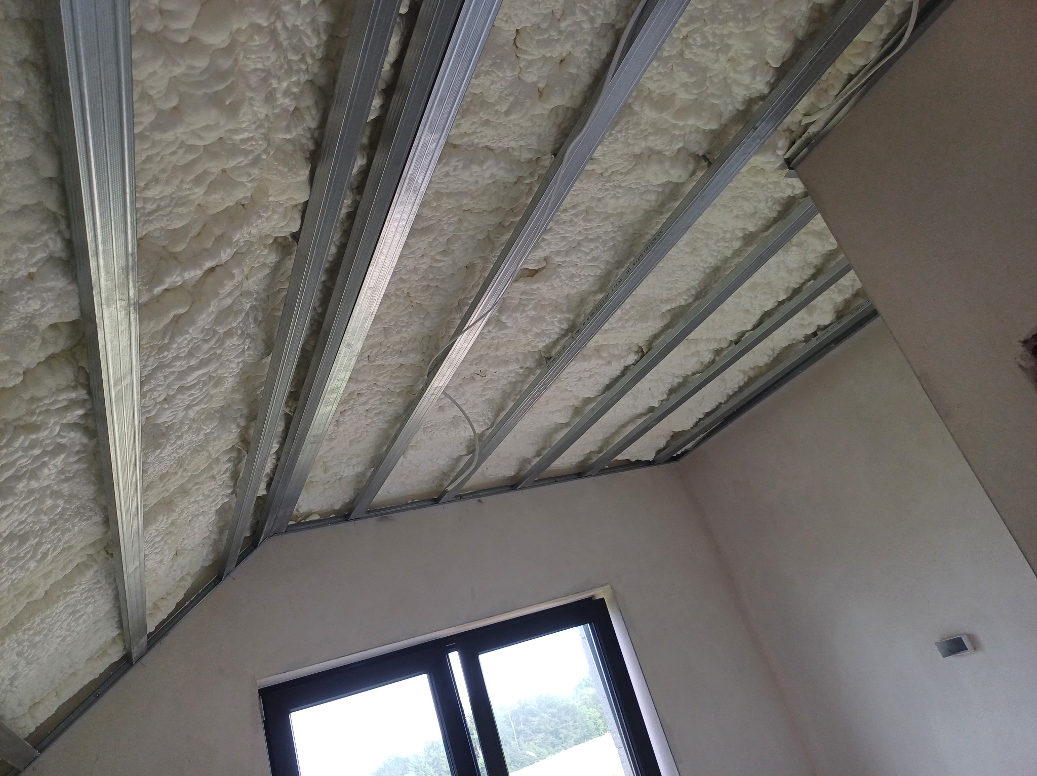 Poddasze z oknem, ocieplone pianą i przygotowane pod montaż płyt gipsowych, widoczne metalowe profile. Skos dachu.