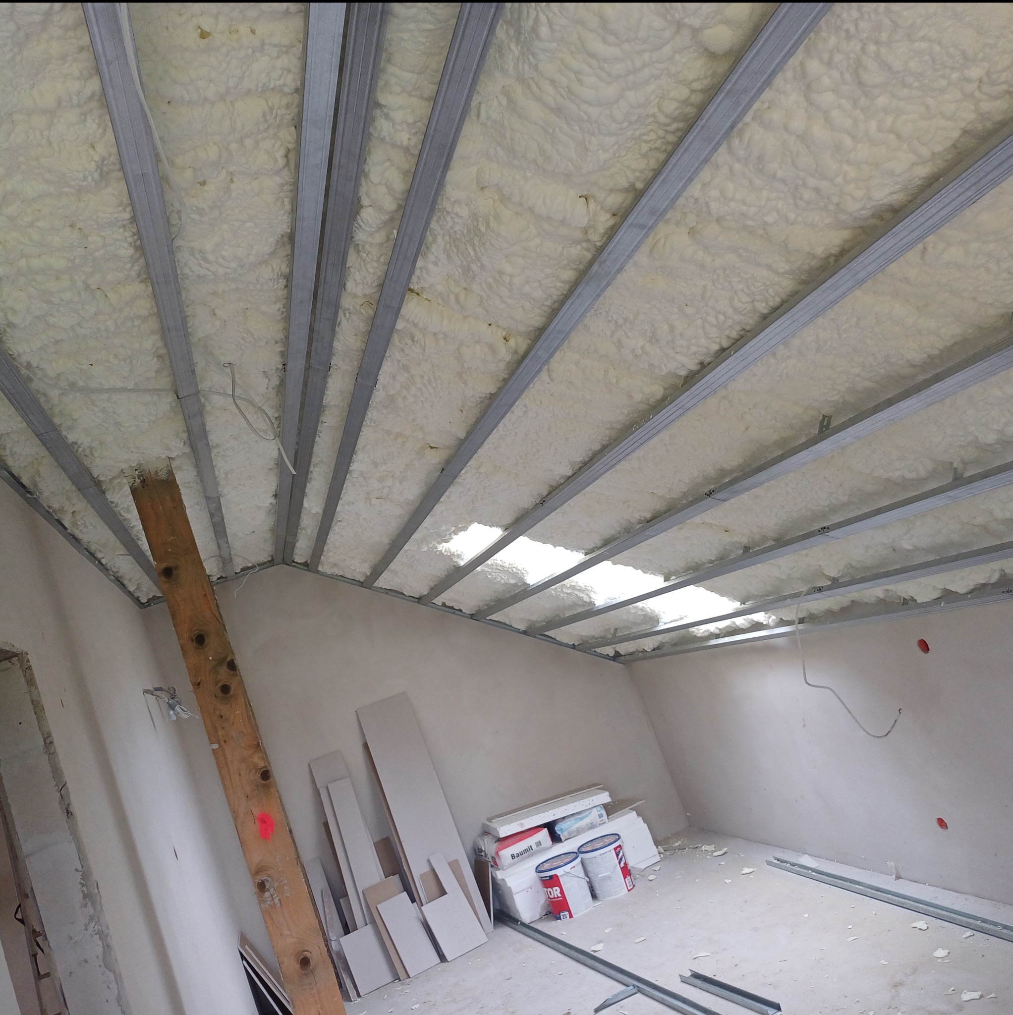 Sufit podwieszany w trakcie izolacji pianą, widoczne profile metalowe i częściowo wypełniona przestrzeń. Na podłodze materiały budowlane i puszki z farbą.