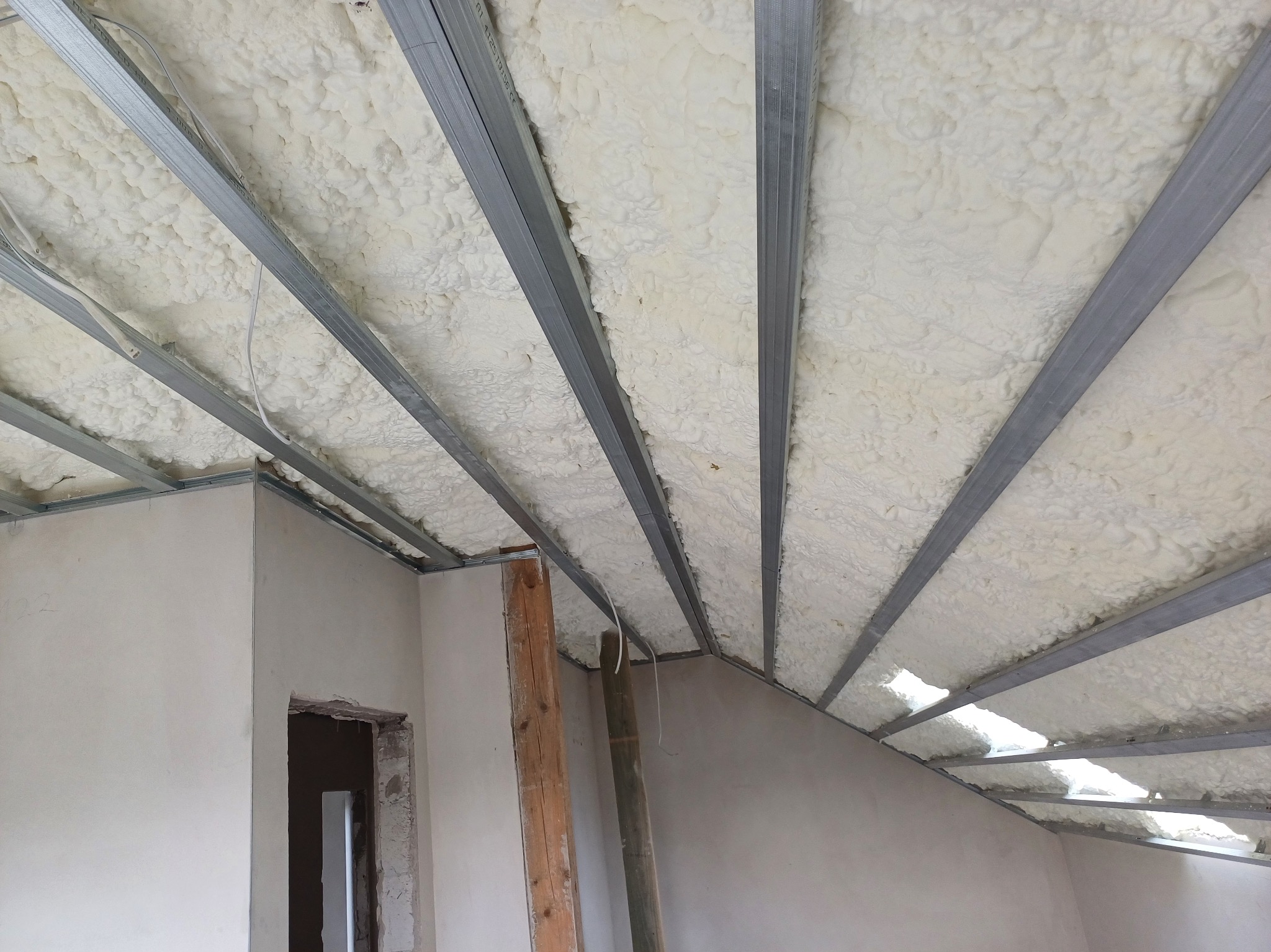 Sufit poddasza z izolacją piankową i metalową konstrukcją nośną, widok z dołu. Fragment ściany z otworem drzwiowym i drewniane podpory.