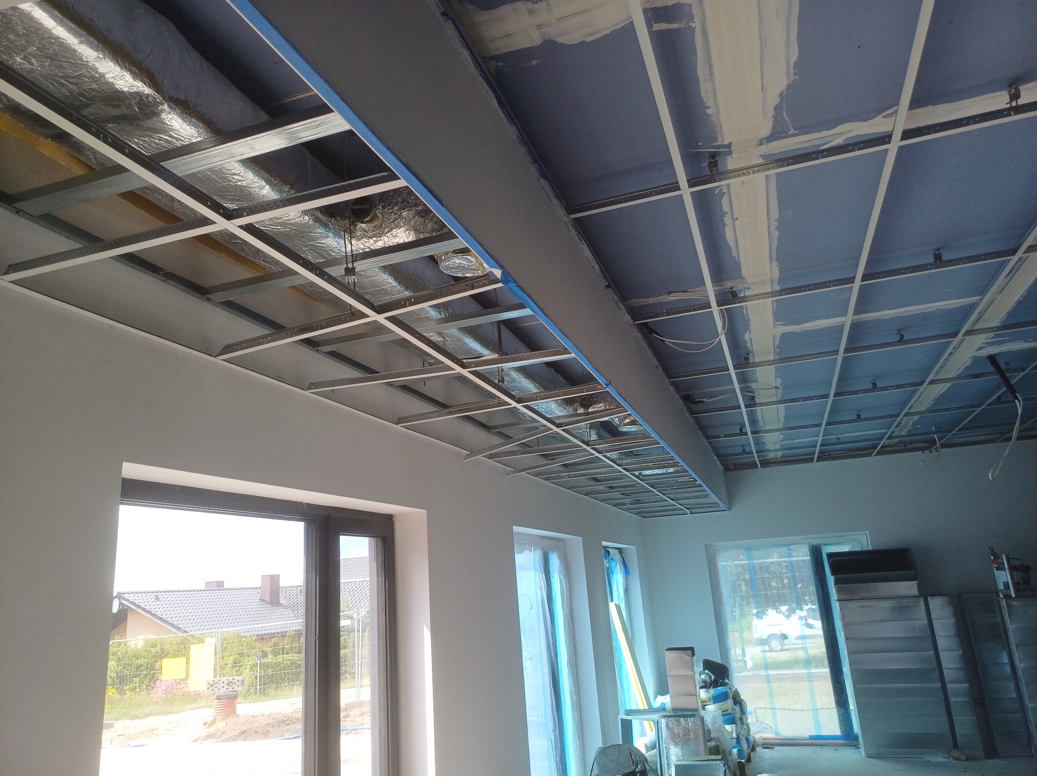 Montaż sufitu podwieszanego z płyt gipsowo-kartonowych, widoczna metalowa konstrukcja i izolacja. Okna z widokiem na dom w tle.