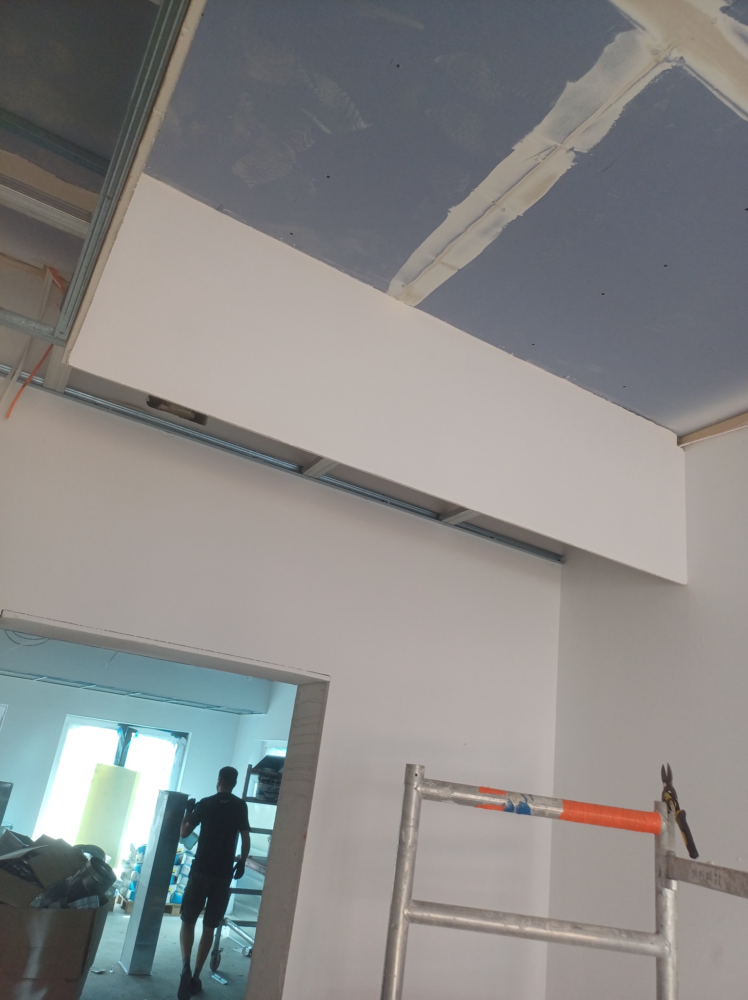 Montaż sufitu podwieszanego z płyty gipsowej, widoczne profile metalowe, szpachlowane łączenia. W tle pracownik i rusztowanie.