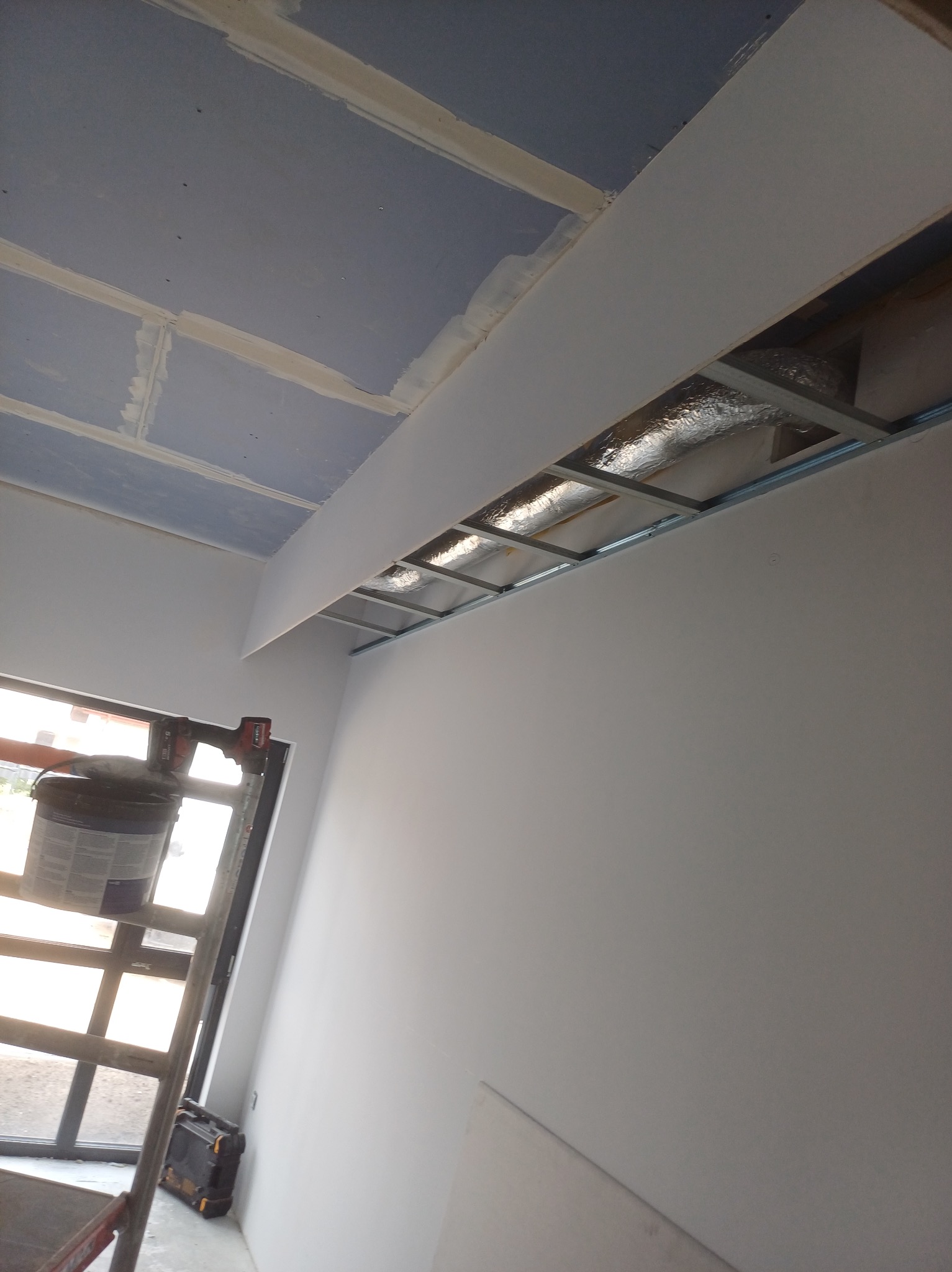 Wykończony fragment sufitu podwieszanego z płyt karton-gips, widok z dołu. Widoczna drabina z narzędziami i wiadro z farbą. Oświetlenie naturalne z okna.
