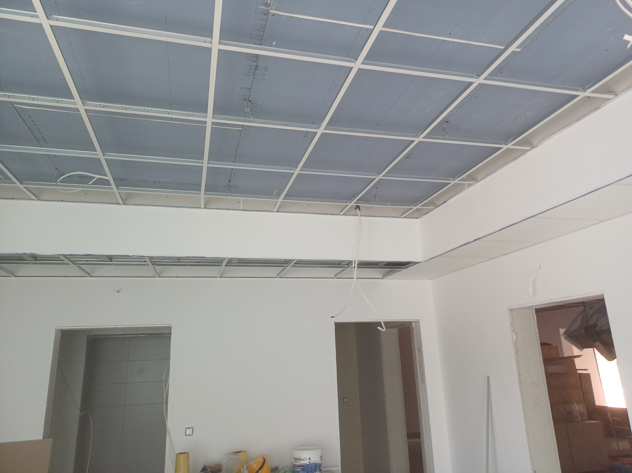 Montaż sufitu podwieszanego z płyt kartonowo-gipsowych na metalowej konstrukcji w jasnym wnętrzu. Widoczne kable i puszki elektryczne.