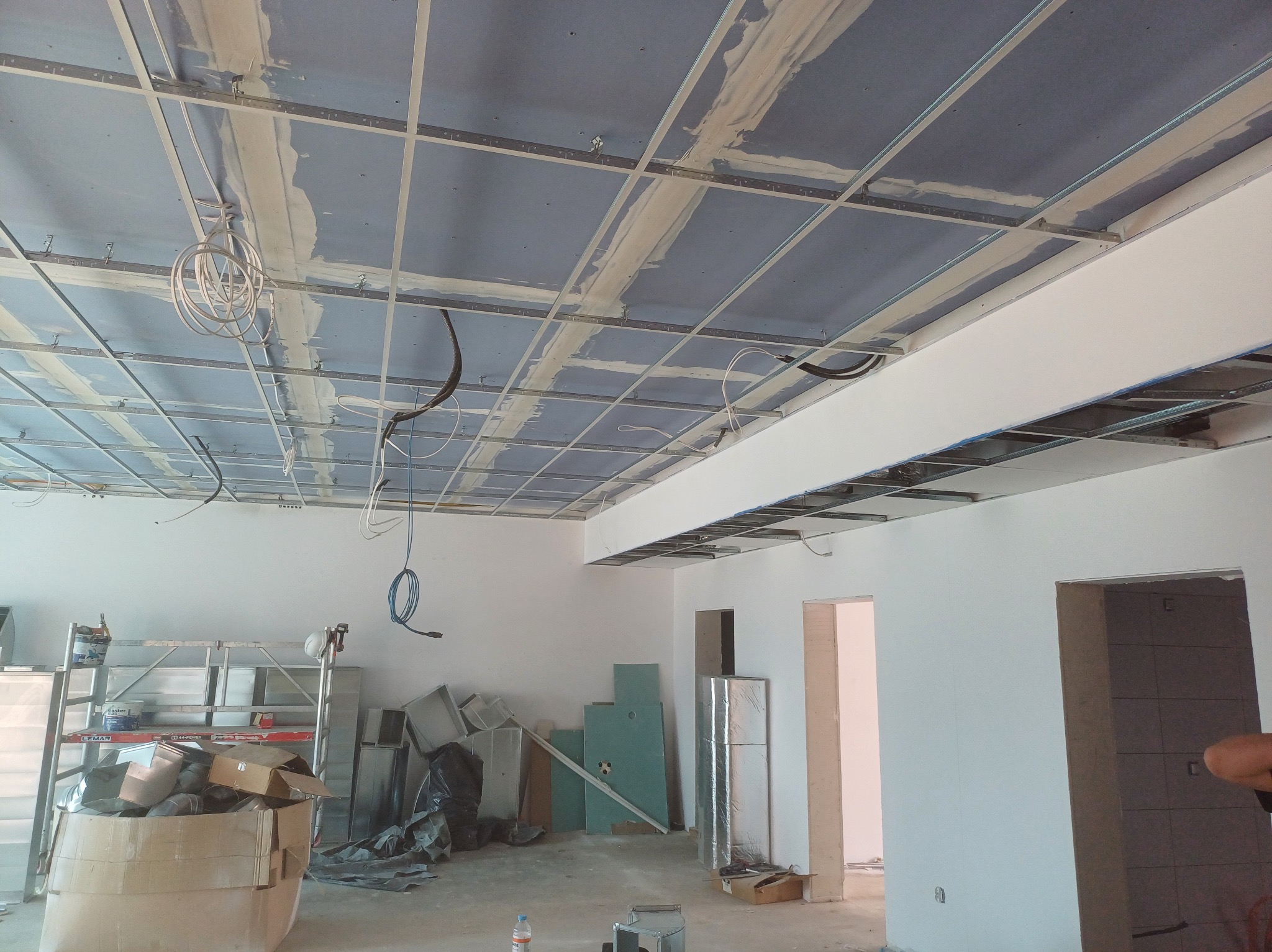 Sufit podwieszany z płyt karton-gips na metalowej konstrukcji, widoczne przewody i elementy instalacji. W tle białe ściany i materiały budowlane.