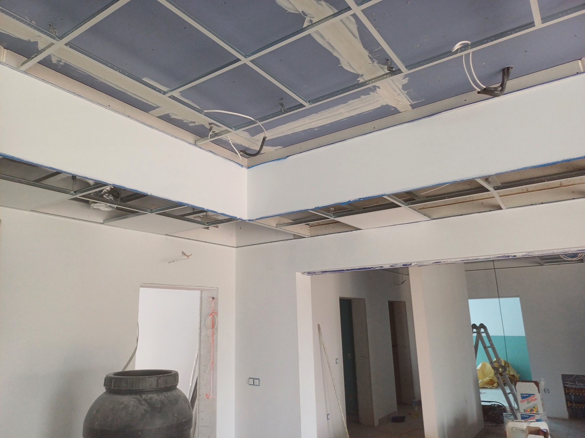 Sufit podwieszany z płyt gipsowo-kartonowych w trakcie montażu, widoczne profile i instalacje. Ściany pomalowane na biało, surowy stan wykończenia wnętrza.