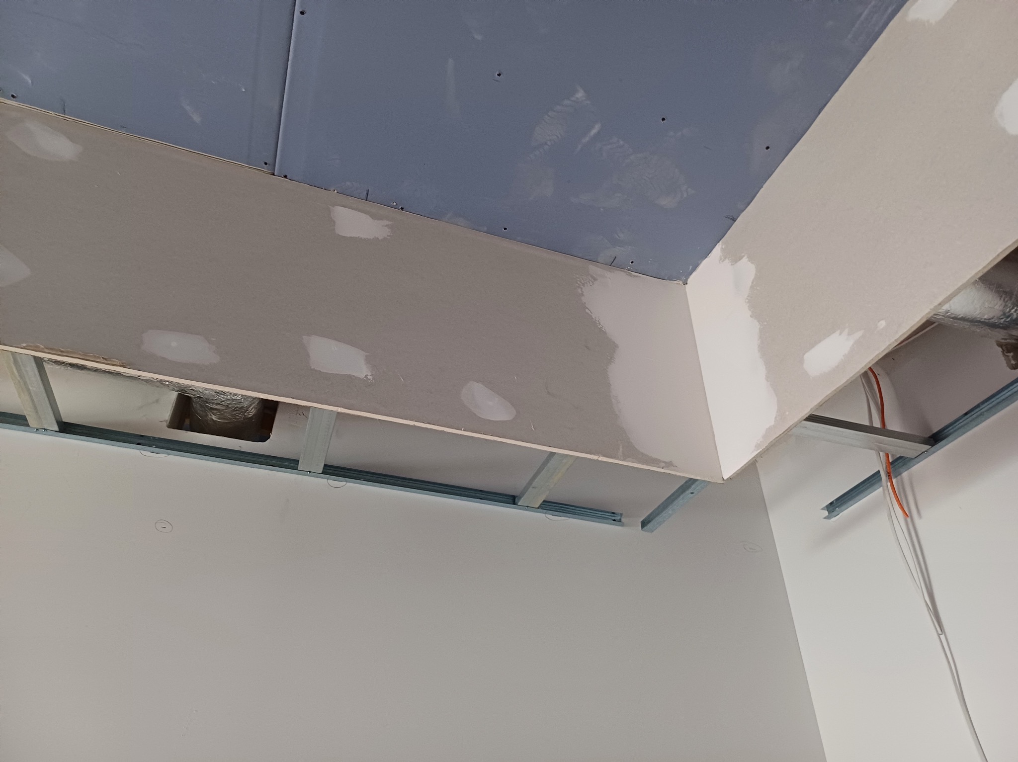 Sufit podwieszany z płyty gipsowej w trakcie szpachlowania, widoczne metalowe profile i fragment instalacji elektrycznej. Narożnik sufitu, jasne ściany.
