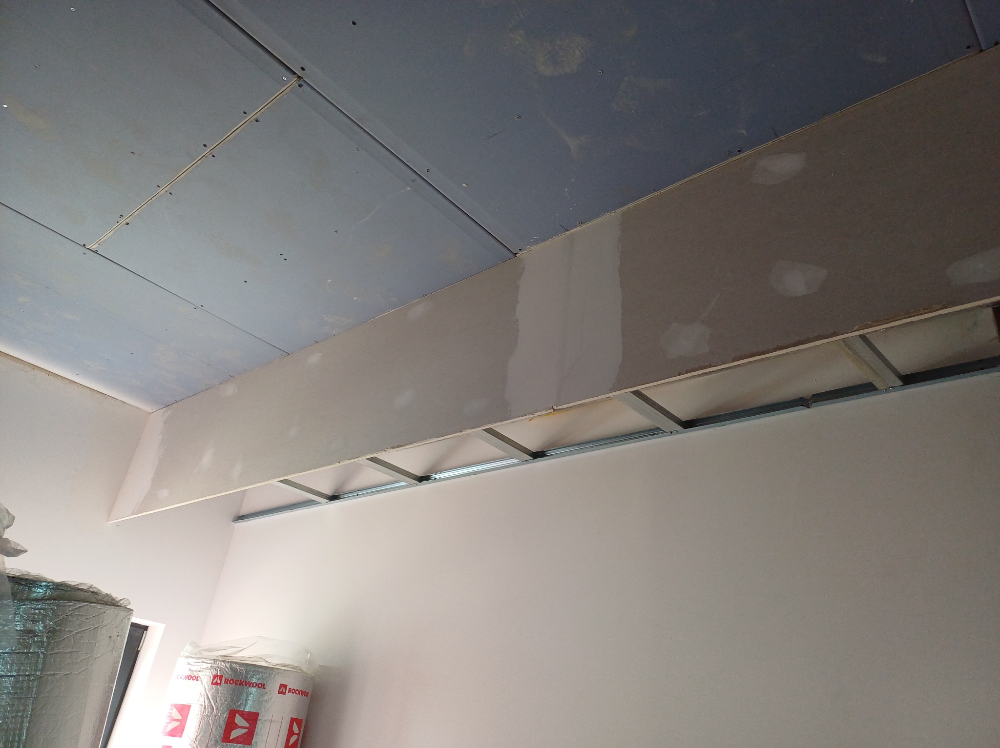 Sufit podwieszany z płyty karton-gips na metalowej konstrukcji, częściowo wyszpachlowany, z widoczną izolacją w tle. Etap prac wykończeniowych.