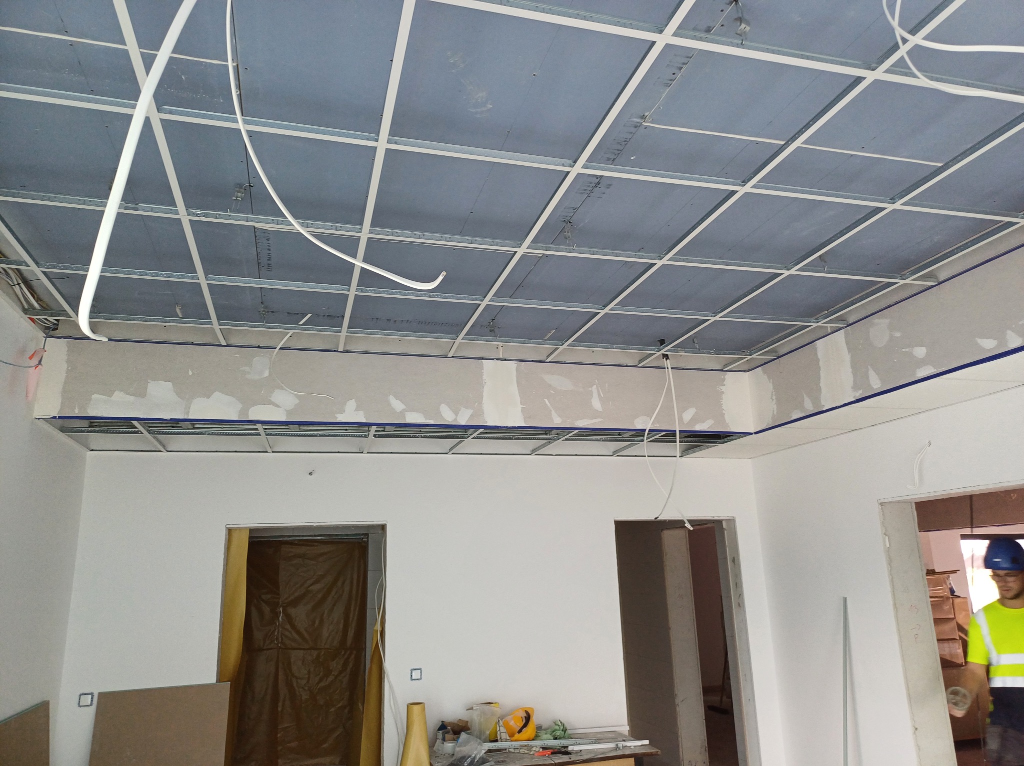 Montaż sufitu podwieszanego z płyt gipsowo-kartonowych na metalowej konstrukcji. Widoczne przewody i częściowo wykończone ściany w jasnym pomieszczeniu.