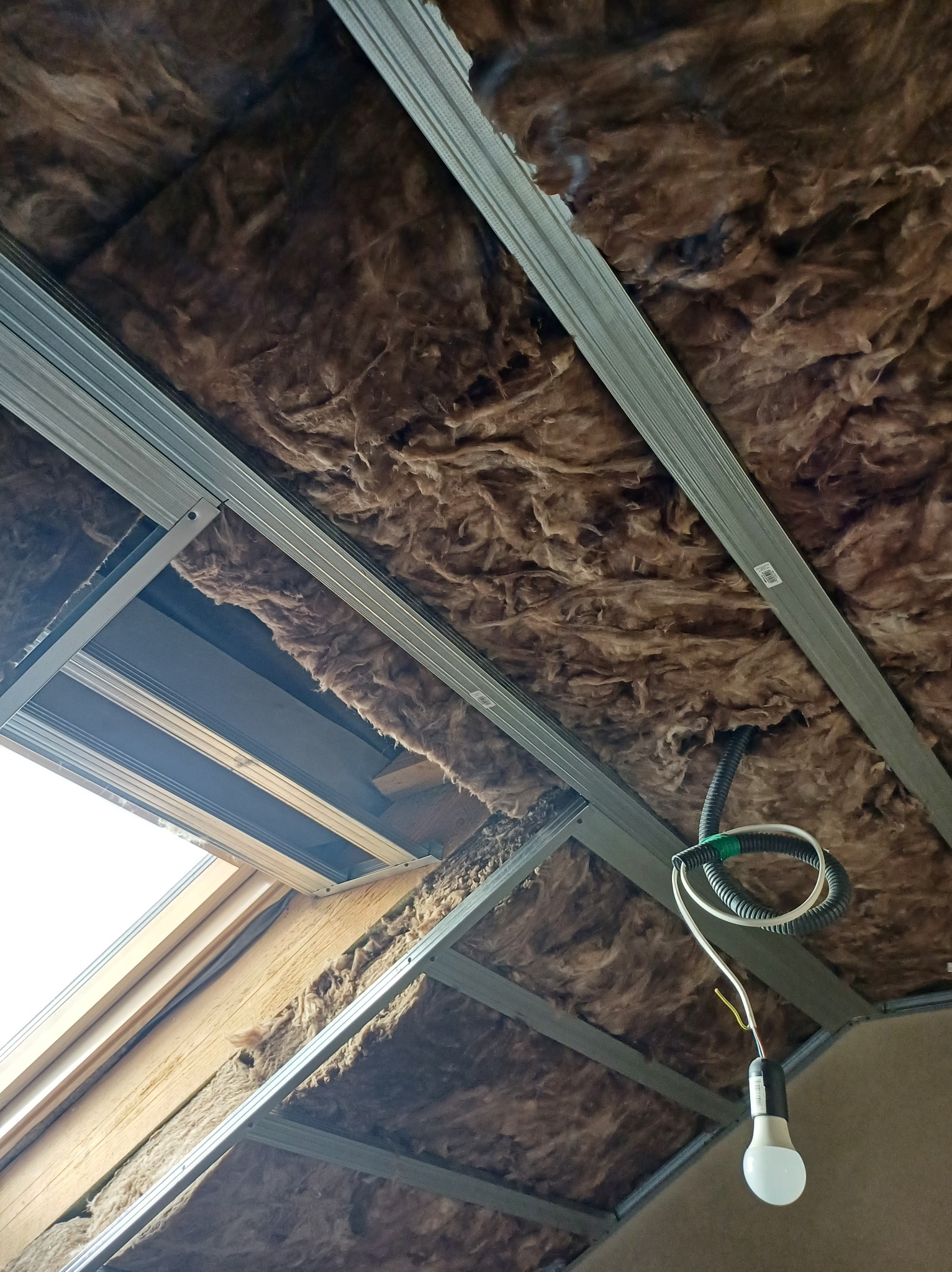 Ocieplenie poddasza wełną mineralną, widoczne metalowe profile konstrukcyjne i okno dachowe. Kabel z żarówką zwisa z sufitu.