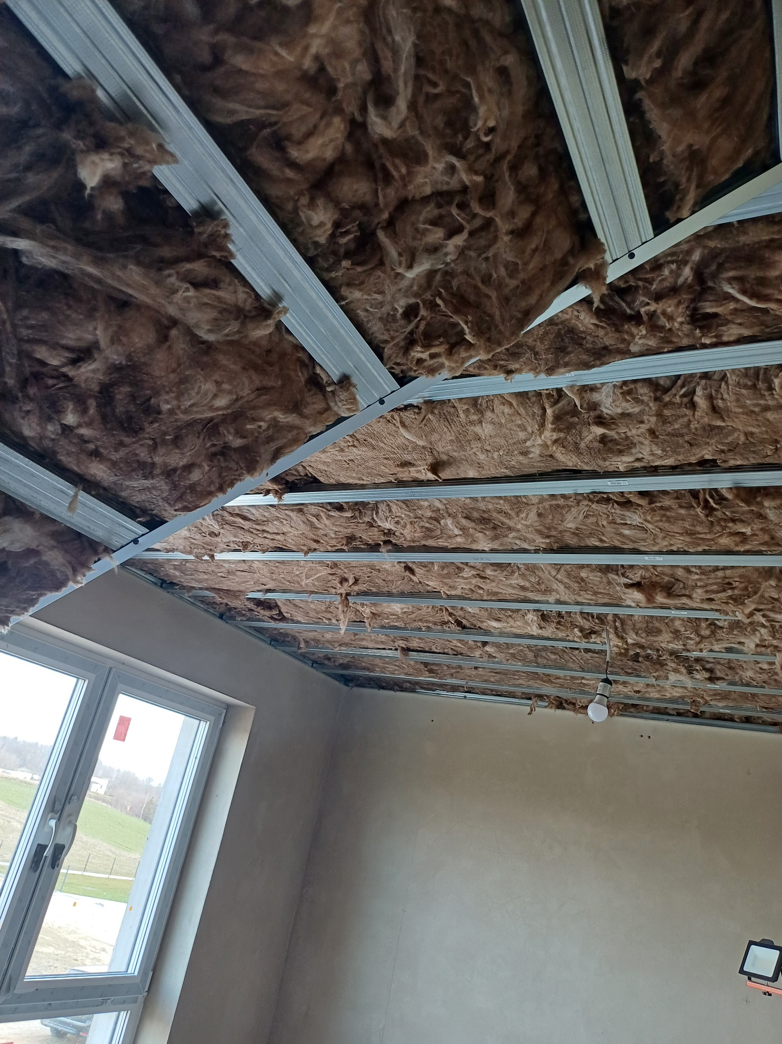 Montaż izolacji z wełny mineralnej na metalowym ruszcie sufitowym w nowym budynku. Widoczne okno i lampa budowlana. Prace wykończeniowe na poddaszu.
