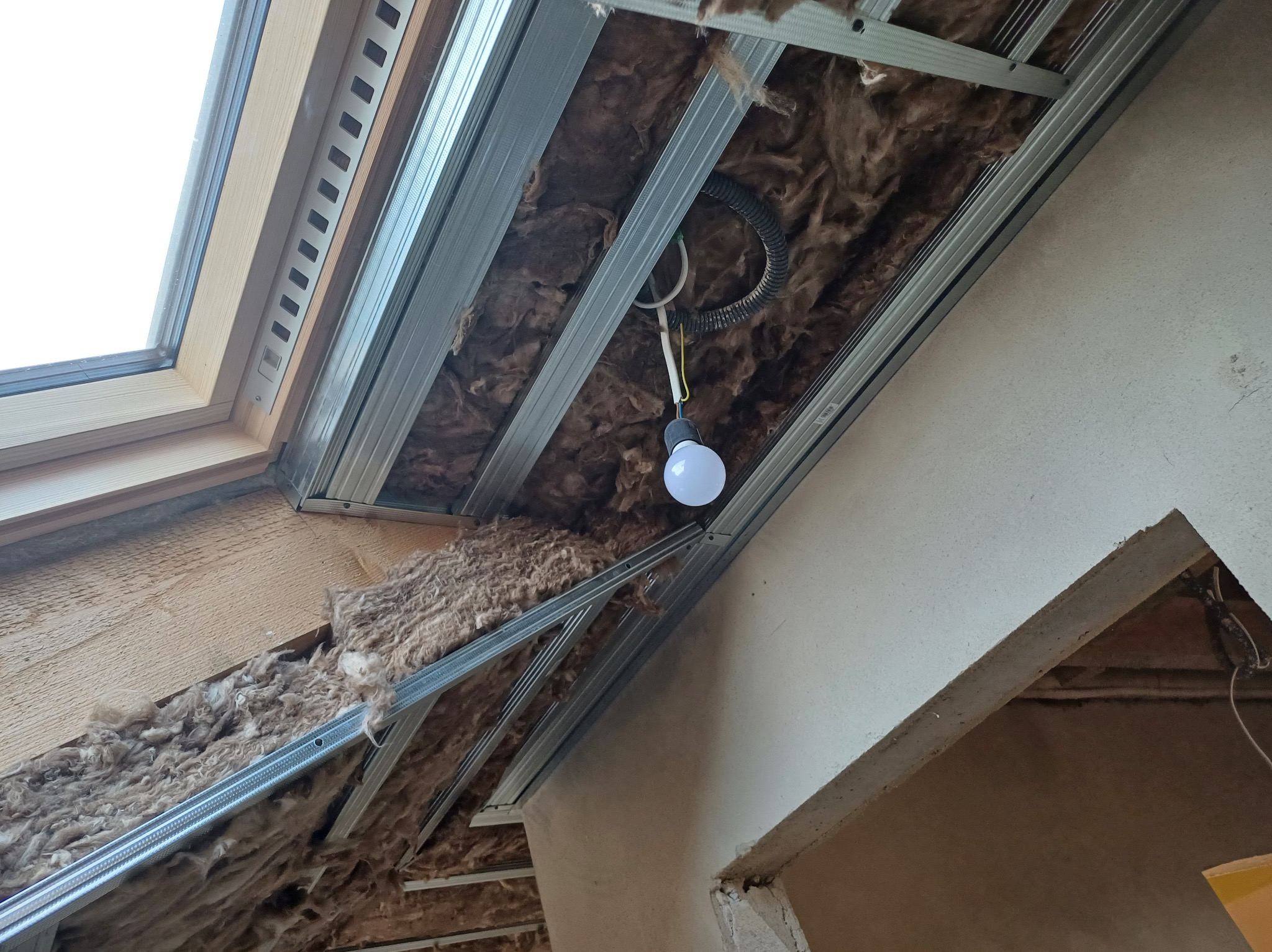 Montaż izolacji z wełny mineralnej i metalowego stelażu pod płytę G-K na poddaszu. Widoczne okno dachowe i prowizoryczne oświetlenie żarówką.