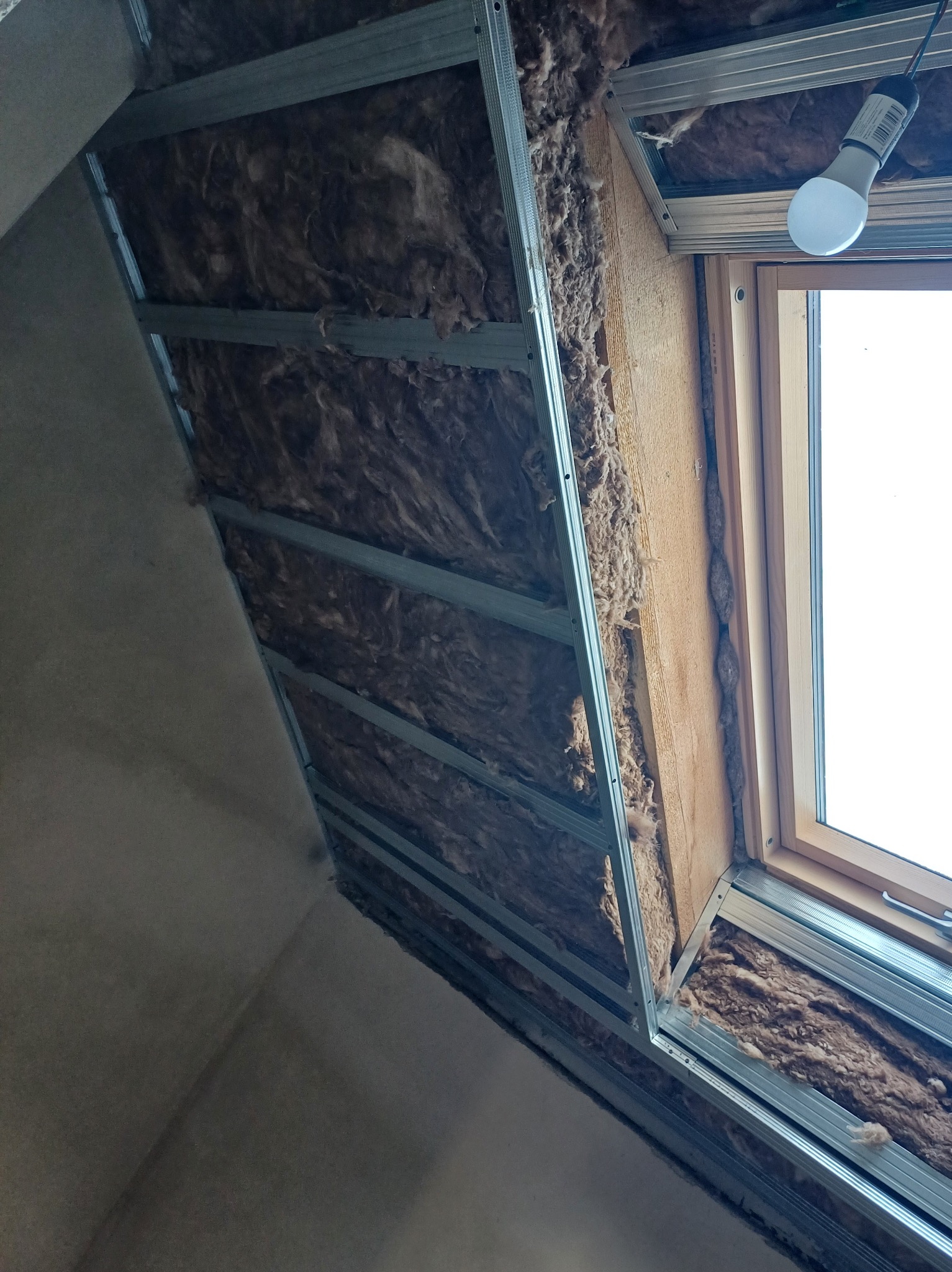 Skośny sufit poddasza z oknem dachowym w trakcie izolacji wełną mineralną, widoczne metalowe profile konstrukcyjne. Ujęcie od dołu, lampa LED w rogu.