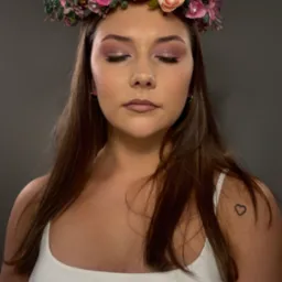 Portret kobiety z zamkniętymi oczami, wiankiem z kwiatów i subtelnym, błyszczącym makijażem oczu. Na ramieniu widoczny tatuaż serca.