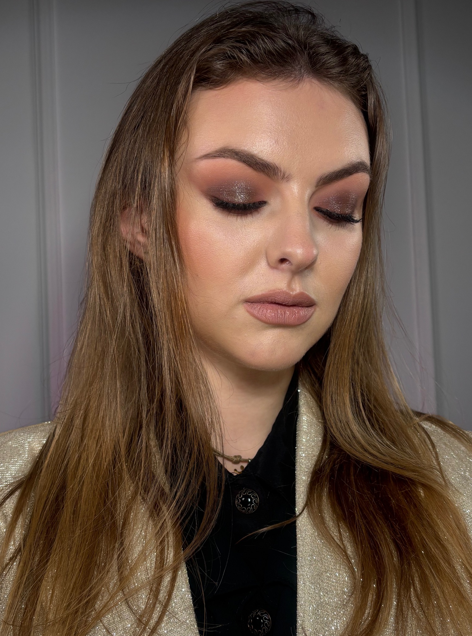 Portret kobiety z brązowym, błyszczącym cieniem na powiekach, beżowymi ustami i delikatnym konturowaniem twarzy, w złotej marynarce.