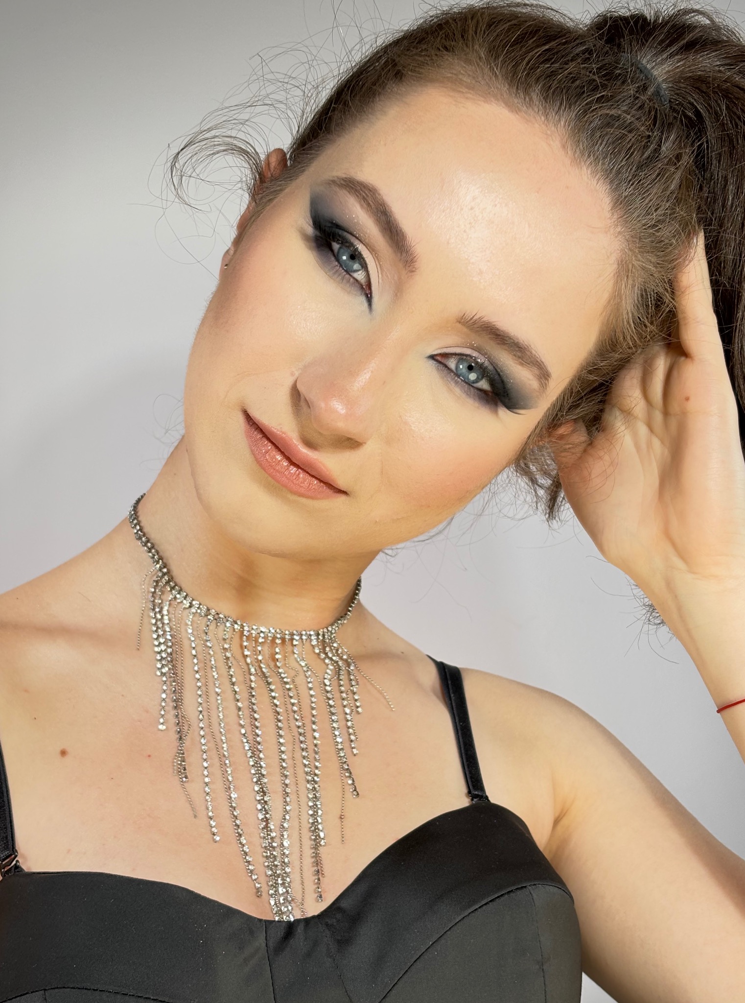 Portret kobiety z intensywnym, wieczorowym make-upem oczu w odcieniach szarości i błyszczącym naszyjnikiem z frędzlami. Uczesanie w wysoki kucyk, delikatny uśmiech.
