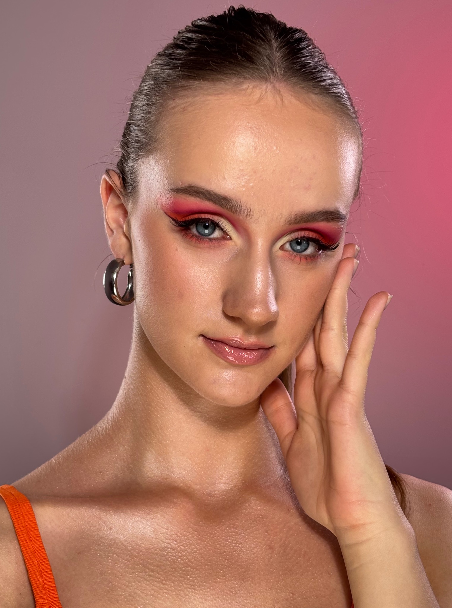 Portret kobiety z intensywnym, czerwono-różowym cieniowaniem powiek i błyszczącymi ustami na tle gradientu różu. Widoczna srebrna kolczyk-obręcz i fragment pomarańczowego ramiączka.