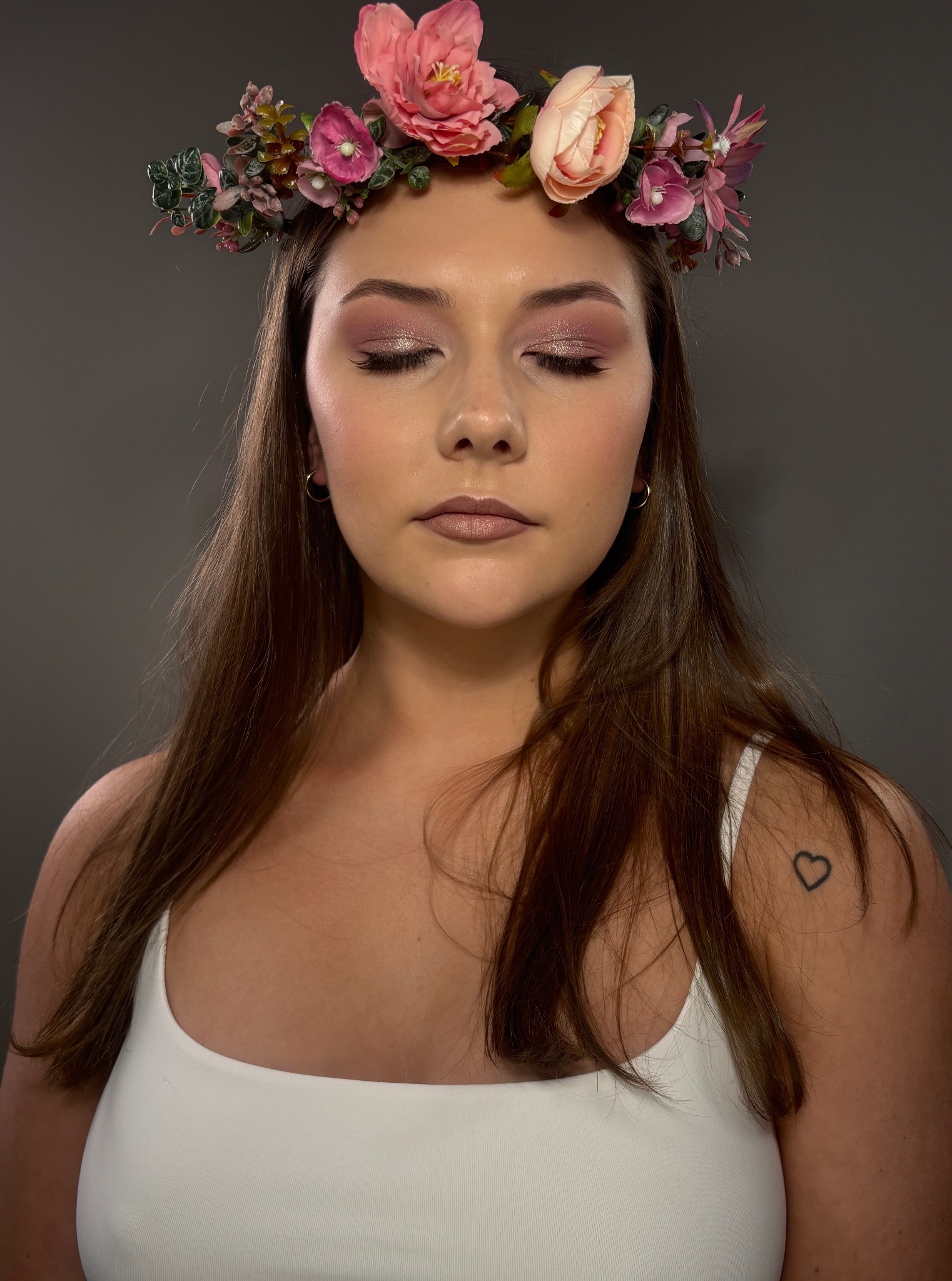 Portret kobiety z zamkniętymi oczami, wiankiem z kwiatów i subtelnym, błyszczącym makijażem oczu. Na ramieniu widoczny tatuaż serca.