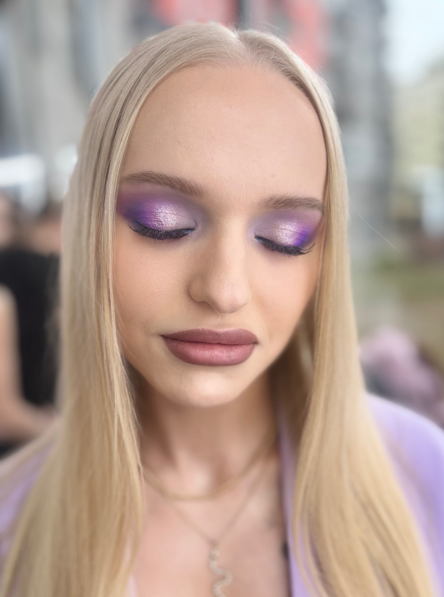 Blondwłosa kobieta z zamkniętymi oczami prezentuje fioletowy, błyszczący make-up oczu i delikatny odcień ust. Widoczny złoty łańcuszek i fioletowa marynarka.