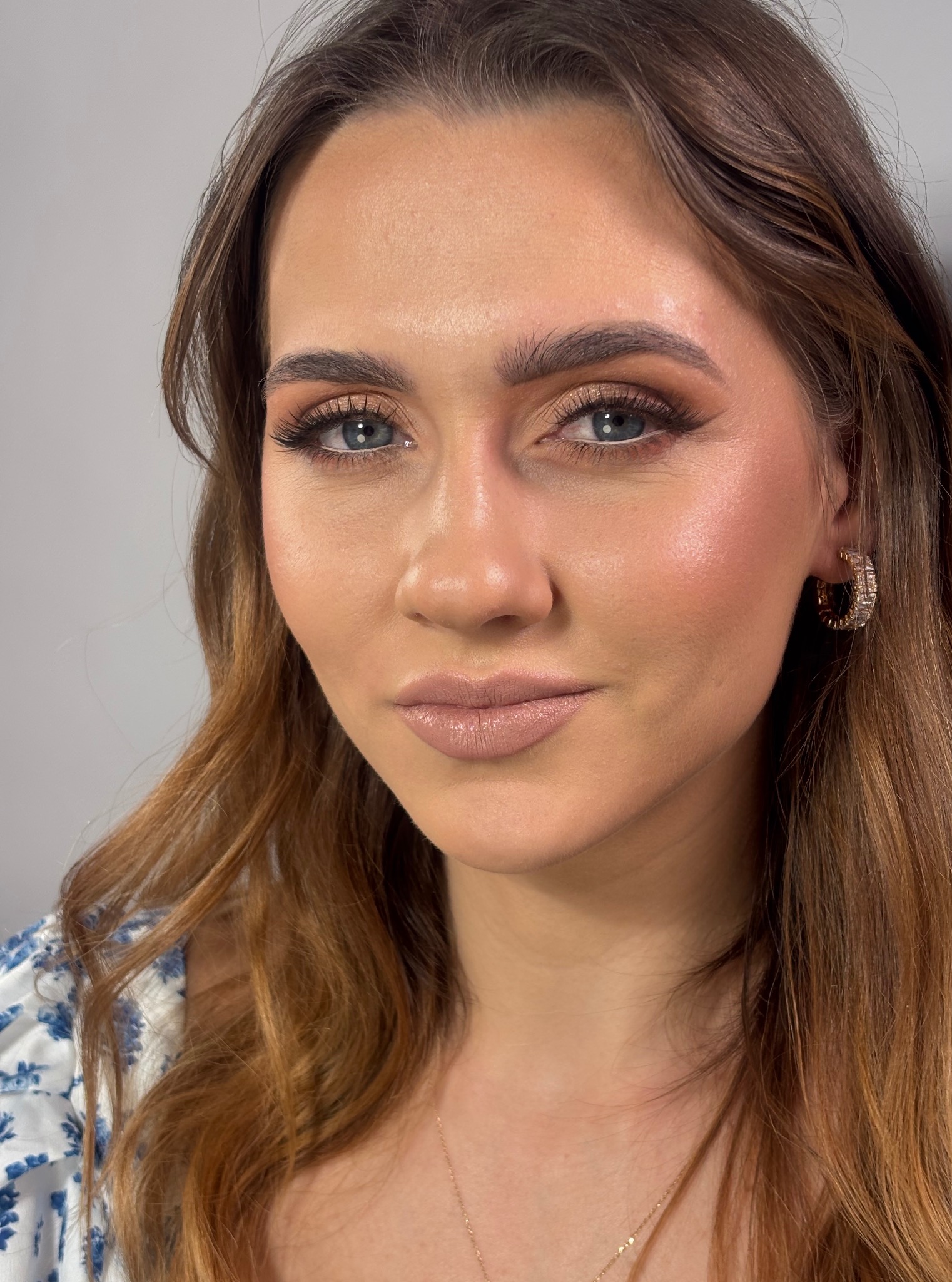 Portret kobiety z subtelnym makijażem, uwydatniającym rysy twarzy i błękitne oczy. Delikatny błyszczyk na ustach i złote kolczyki dodają elegancji.