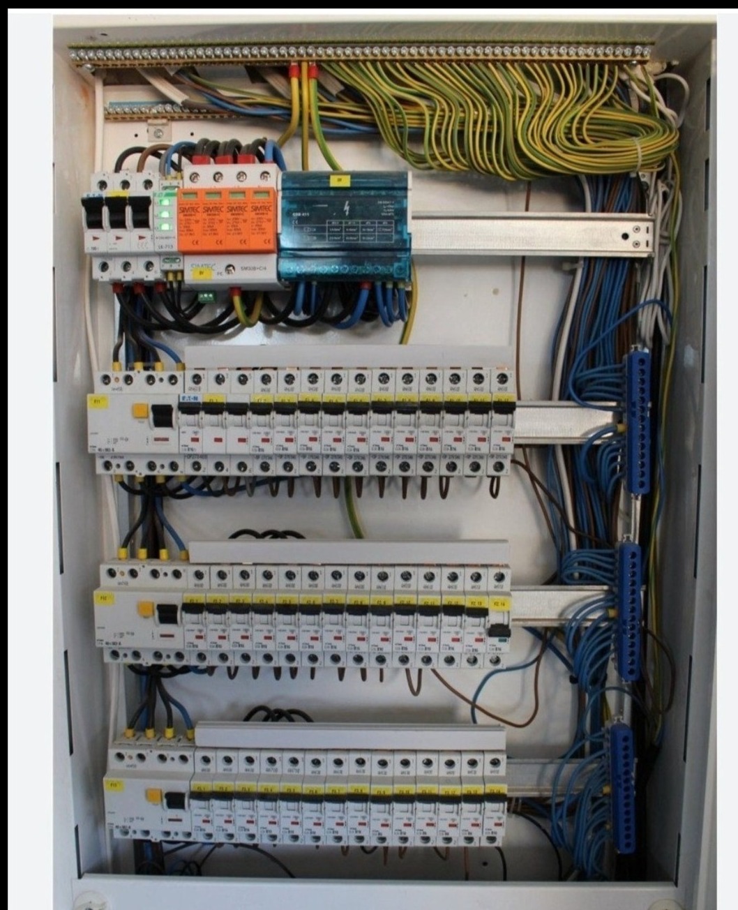 Rozdzielnia elektryczna z bezpiecznikami, okablowaniem i zabezpieczeniami przepięciowymi. Widoczne elementy Simetec i liczne przewody w kolorach żółtym, niebieskim i brązowym.