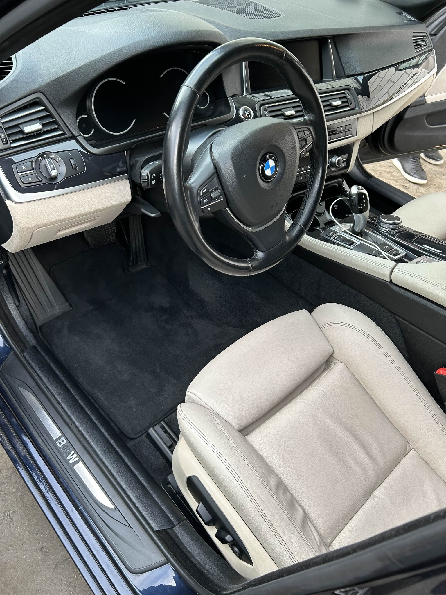 Wnętrze eleganckiego BMW serii 5 z jasną skórzaną tapicerką i czarnymi dywanikami. Widoczna deska rozdzielcza, kierownica z logo BMW i automatyczna skrzynia biegów.