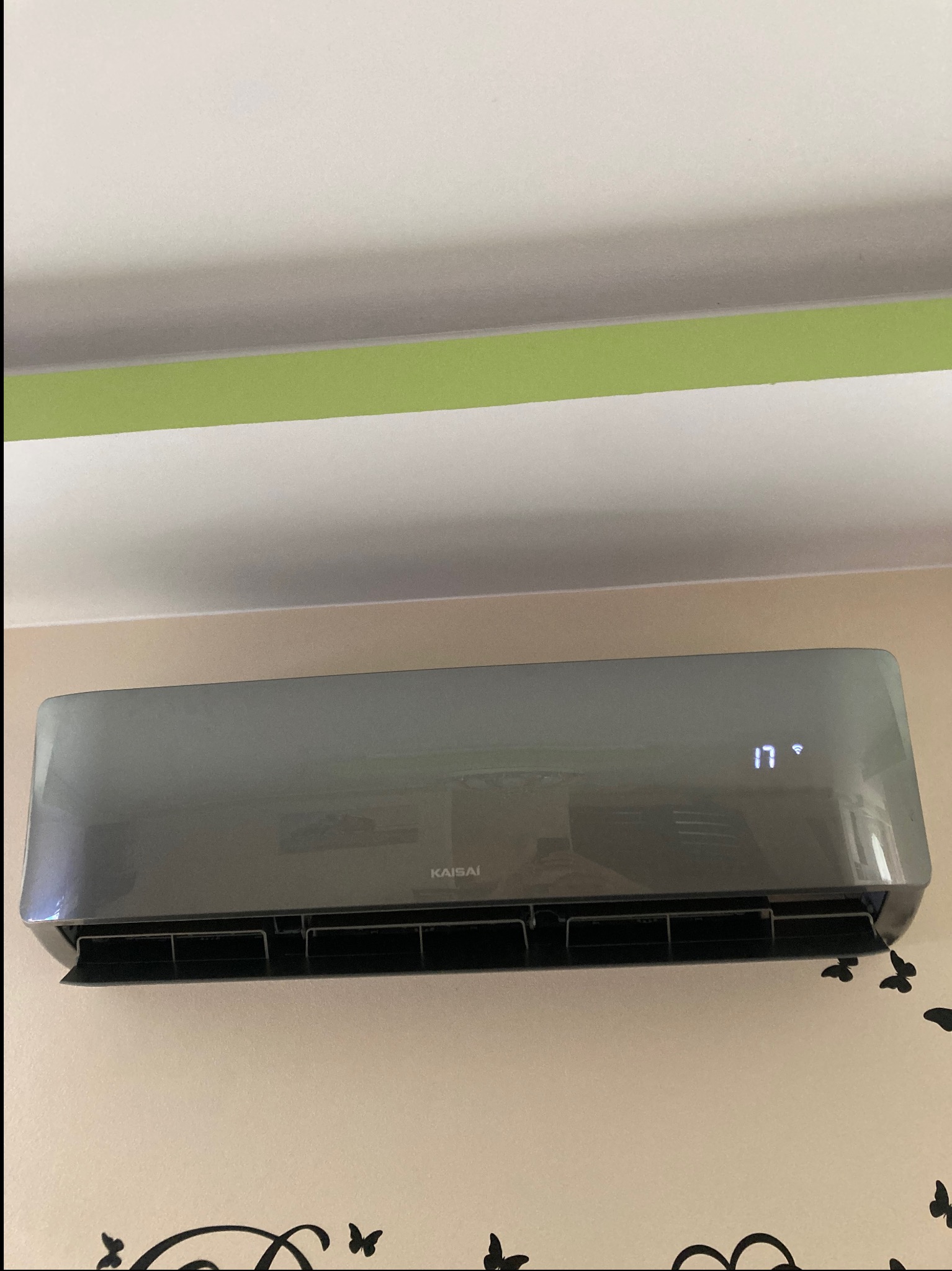 Szary klimatyzator Kaisai na ścianie z dekoracyjnym wzorem motyli, wyświetlający temperaturę 17 stopni. Widoczny fragment sufitu z zielonym paskiem.