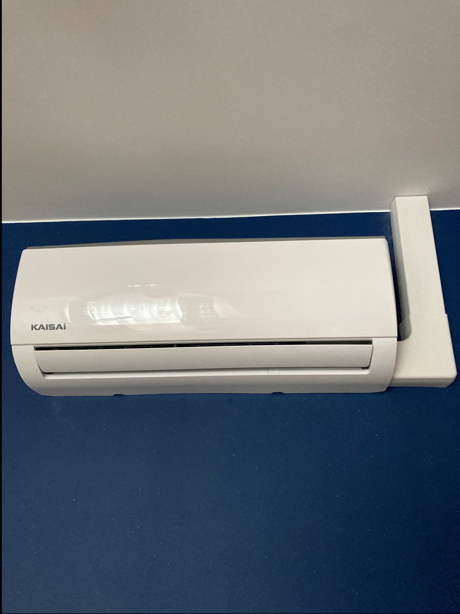 Biała klimatyzacja Kaisai zamontowana na granatowej ścianie, widok z dołu. Widoczne mocowanie klimatyzatora do ściany. Szare tło sufitu.