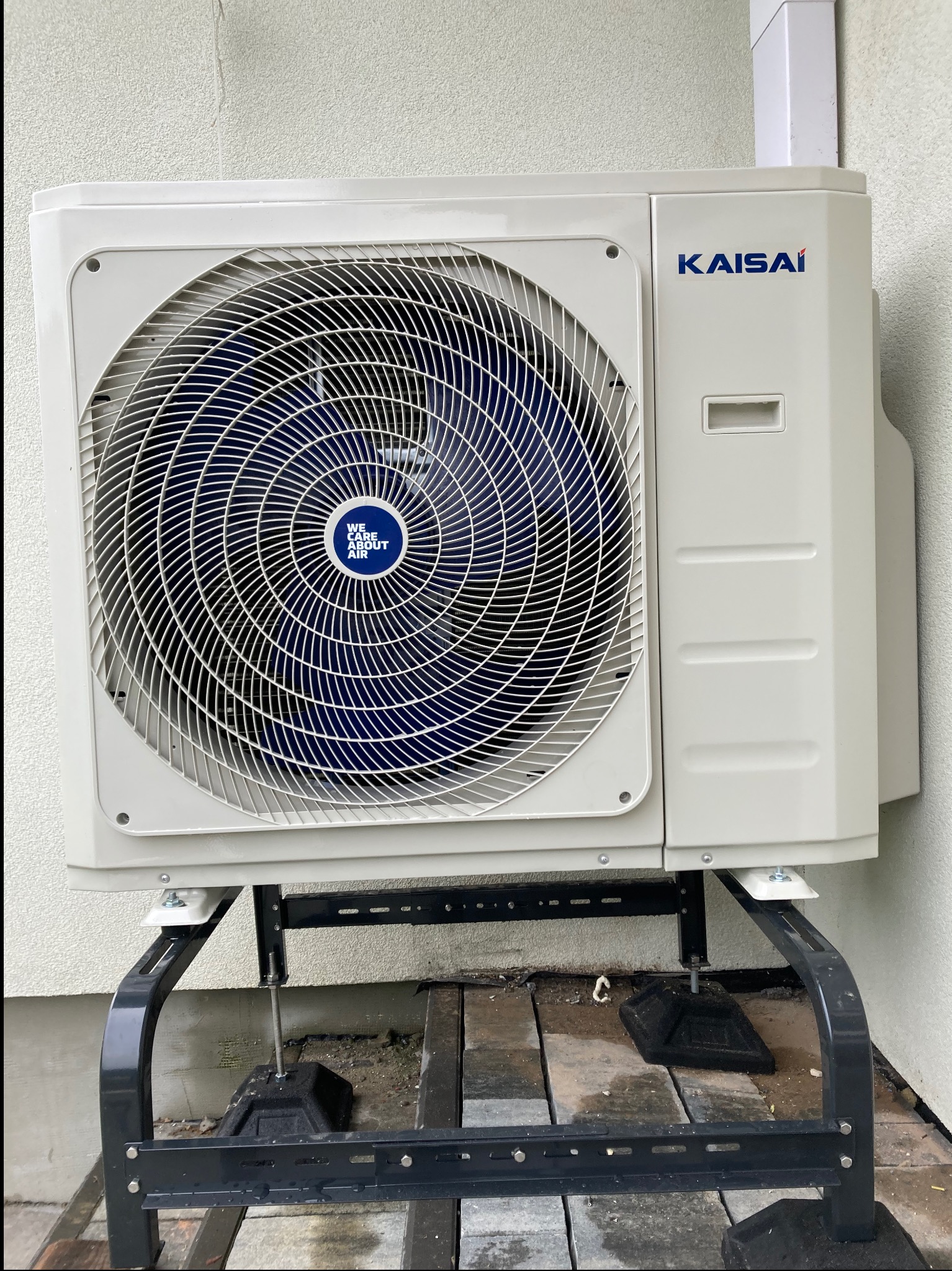 Kompaktowa, zewnętrzna jednostka klimatyzacji Kaisai, zamontowana na czarnej, metalowej podstawie przy ścianie budynku o jasnej elewacji. Widoczne detale konstrukcyjne.