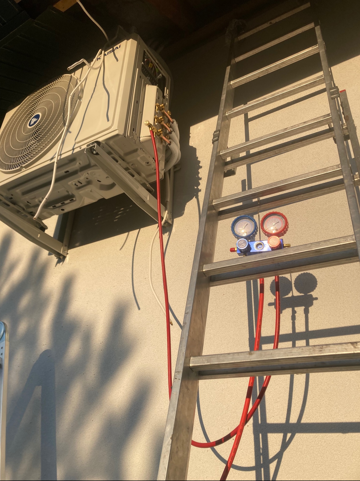 Montaż klimatyzatora Kaisai na elewacji budynku. Widoczna drabina, manometry i przewody chłodnicze. Oświetlenie boczne podkreśla fakturę ściany.