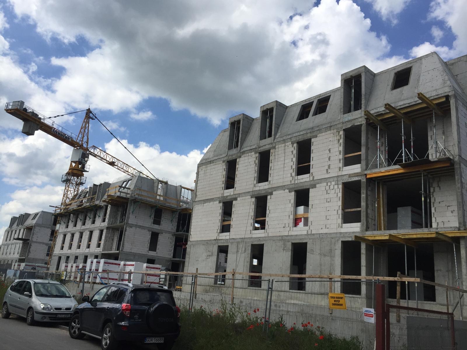 Wznoszenie budynków wielorodzinnych w Serocku. Widoczny żuraw budowlany i surowy stan budynków z białego bloczka, z częściowo zamontowanymi rusztowaniami.