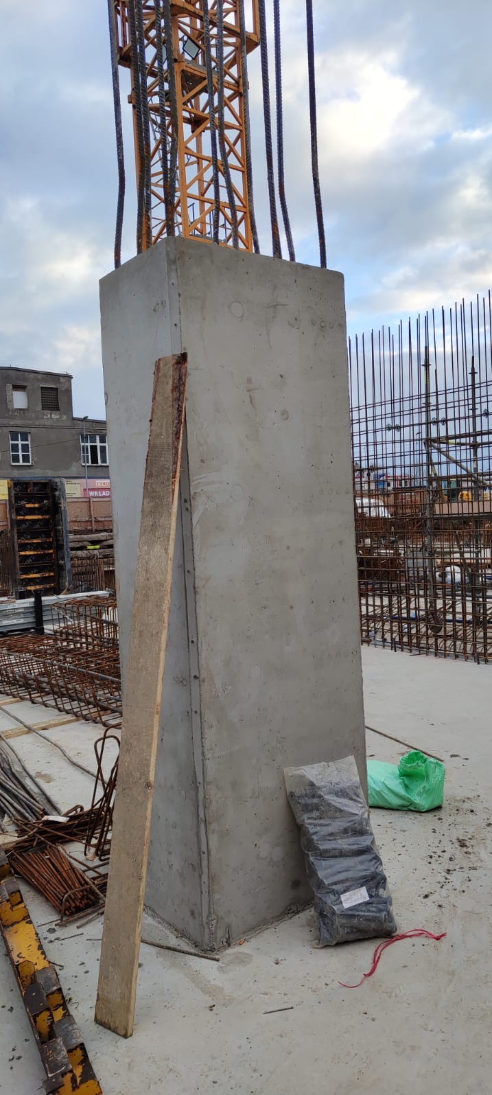 Żelbetowy słup konstrukcyjny z wystającymi prętami zbrojeniowymi, widoczne elementy szalunku i zbrojenia na placu budowy. Częściowo widoczny żuraw wieżowy.