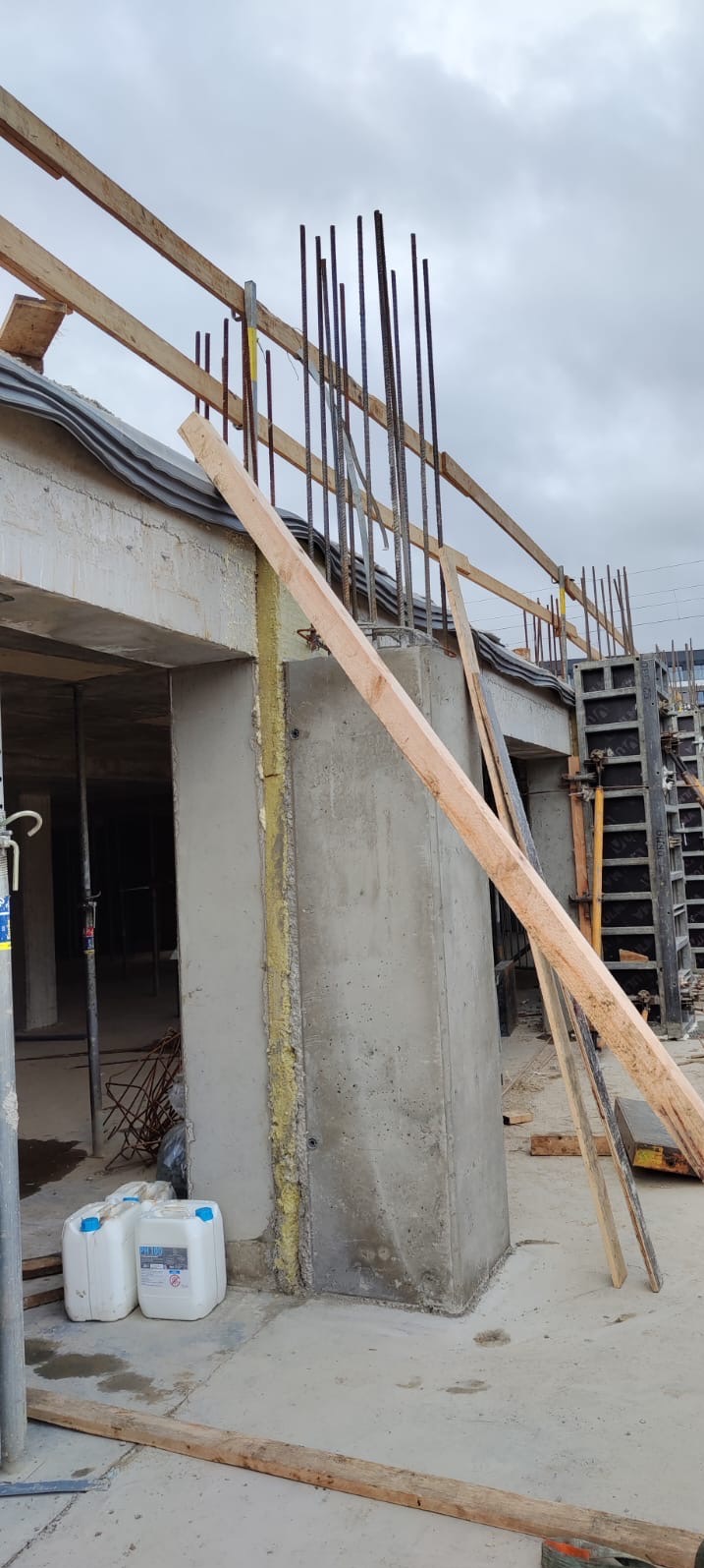 Betonowy szkielet budynku w trakcie budowy. Widoczne zbrojenia, szalunki i materiały izolacyjne. Częściowo zadaszona konstrukcja w stanie surowym otwartym.