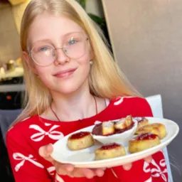 Uśmiechnięta dziewczyna w czerwonej bluzce prezentuje talerz z mini serniczkami z dżemem i sosem w białej miseczce, idealne na słodki catering.