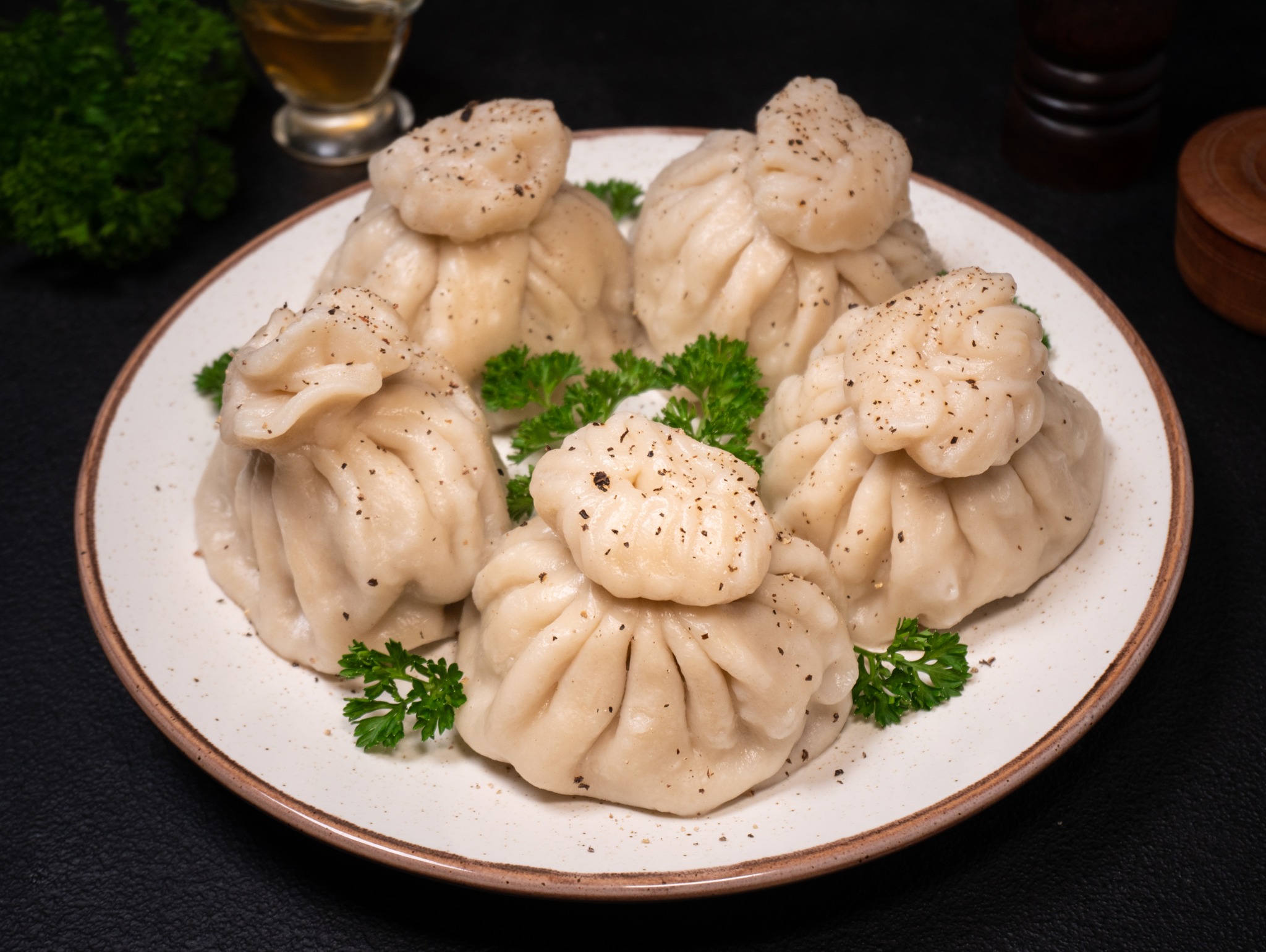 Bliski plan gruzińskich pierogów chinkali na talerzu z dekoracją z zielonej pietruszki, posypanych pieprzem, na ciemnym tle. Widoczny kieliszek i drewniane naczynie.