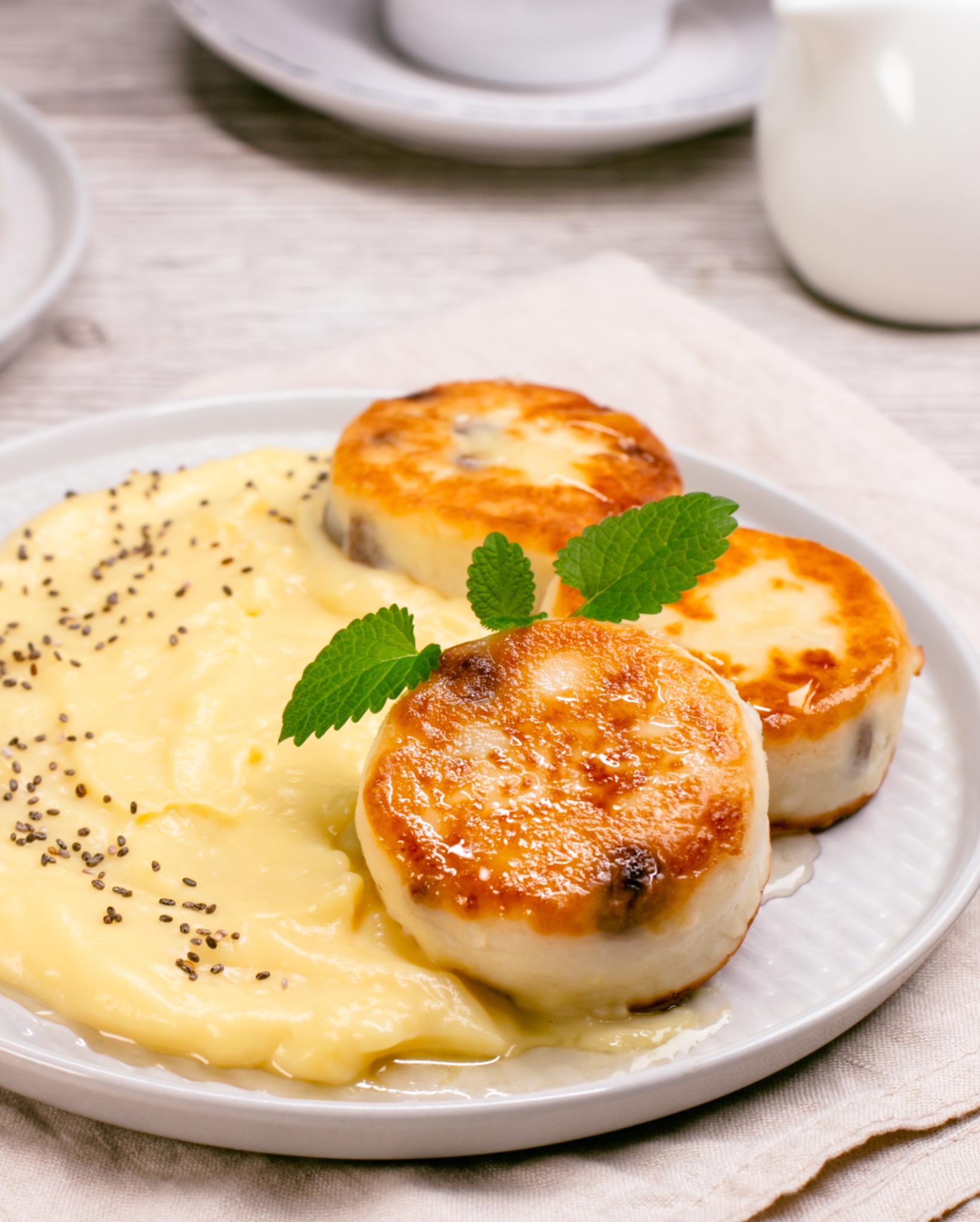 Delikatne, złociste serniczki na talerzu z kremowym sosem i świeżą miętą. Jasne, naturalne światło podkreśla teksturę potrawy. Elegancka kompozycja kulinarna.