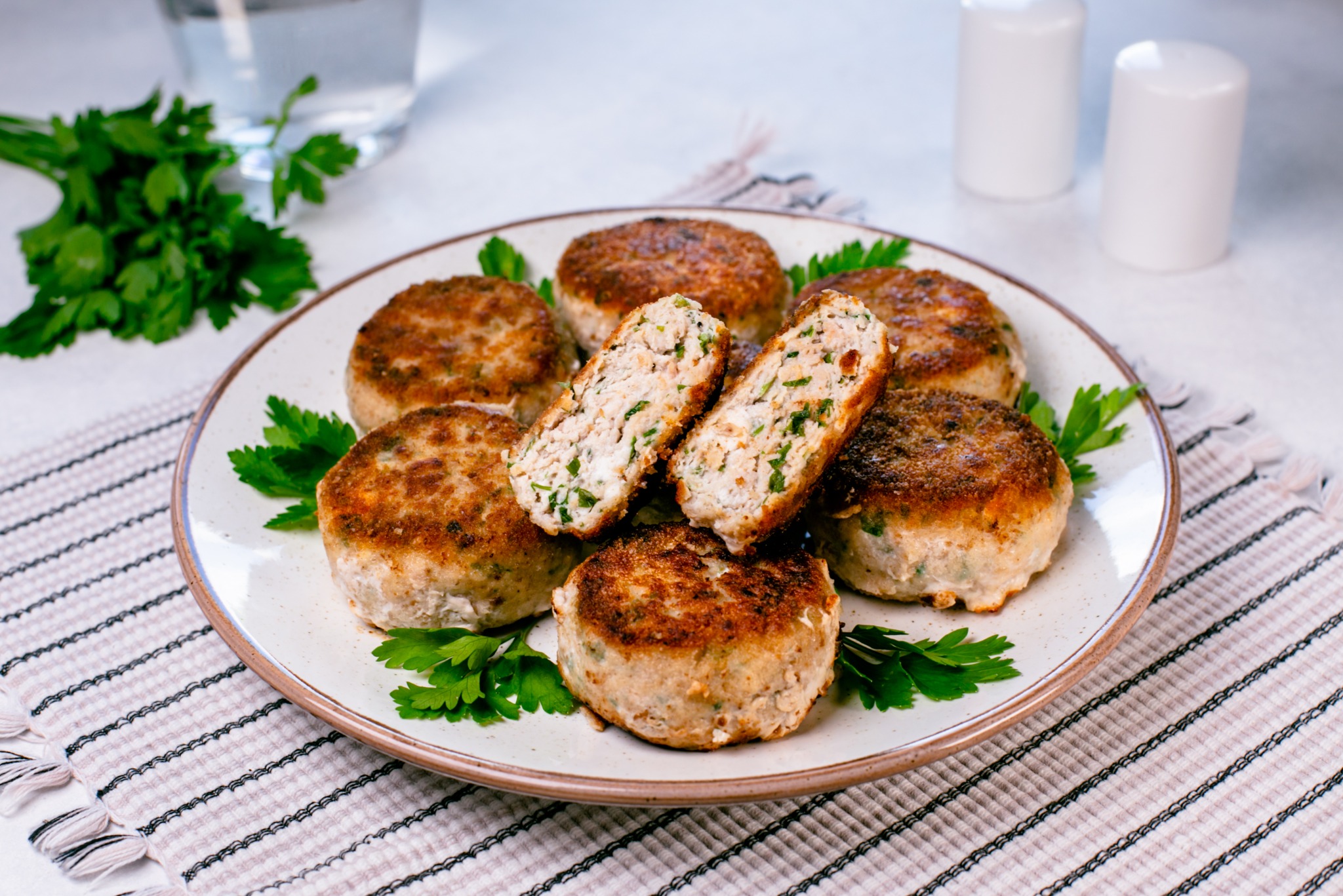 Apetyczne kotleciki drobiowe na talerzu, udekorowane świeżą pietruszką, ustawione na paskowanym ręczniku. W tle szklanka wody i przyprawniki.