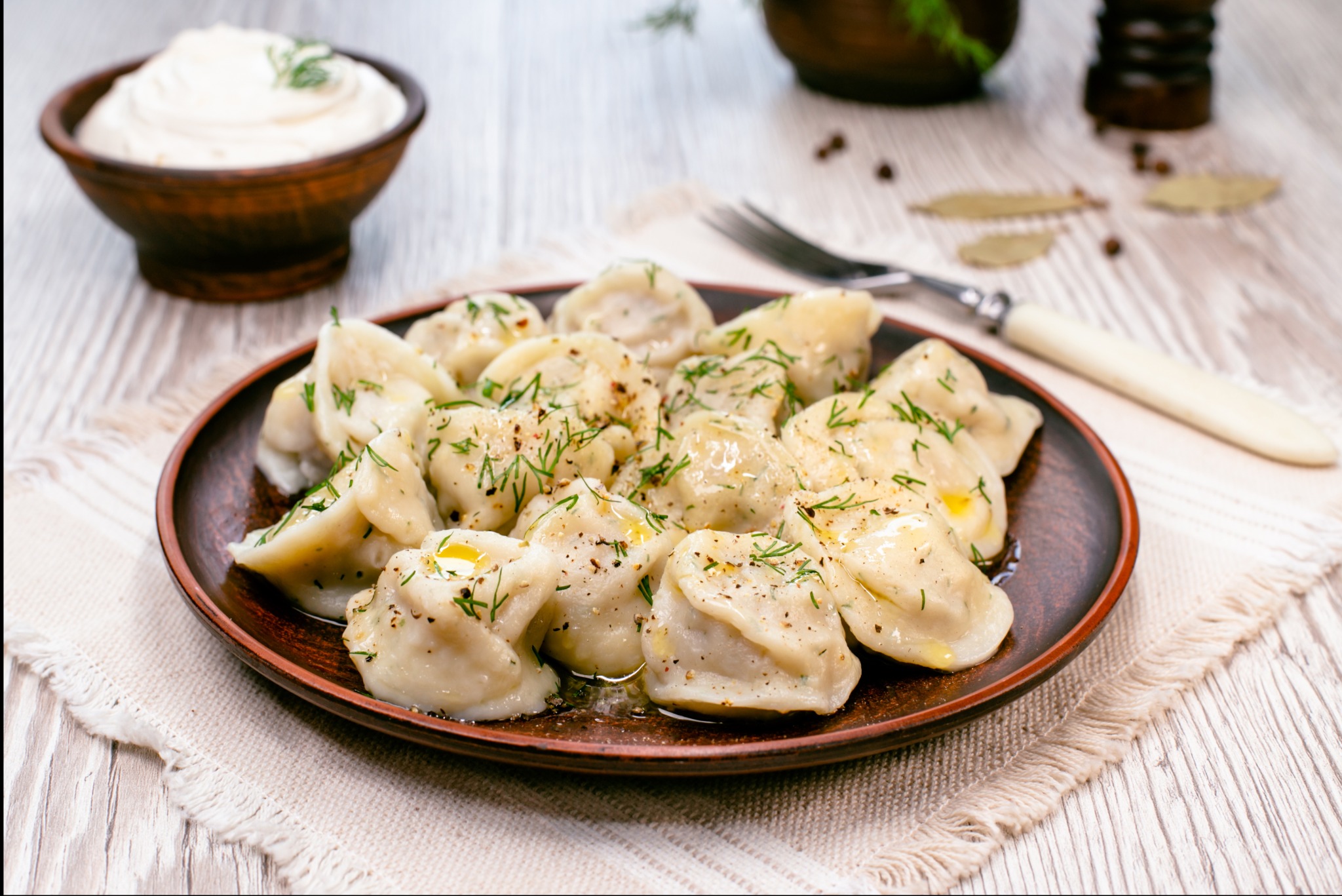 Ręcznie robione pierogi na brązowym talerzu, skropione masłem i posypane koperkiem, obok sos i sztućce na tle drewnianego stołu i lnianej serwetki.