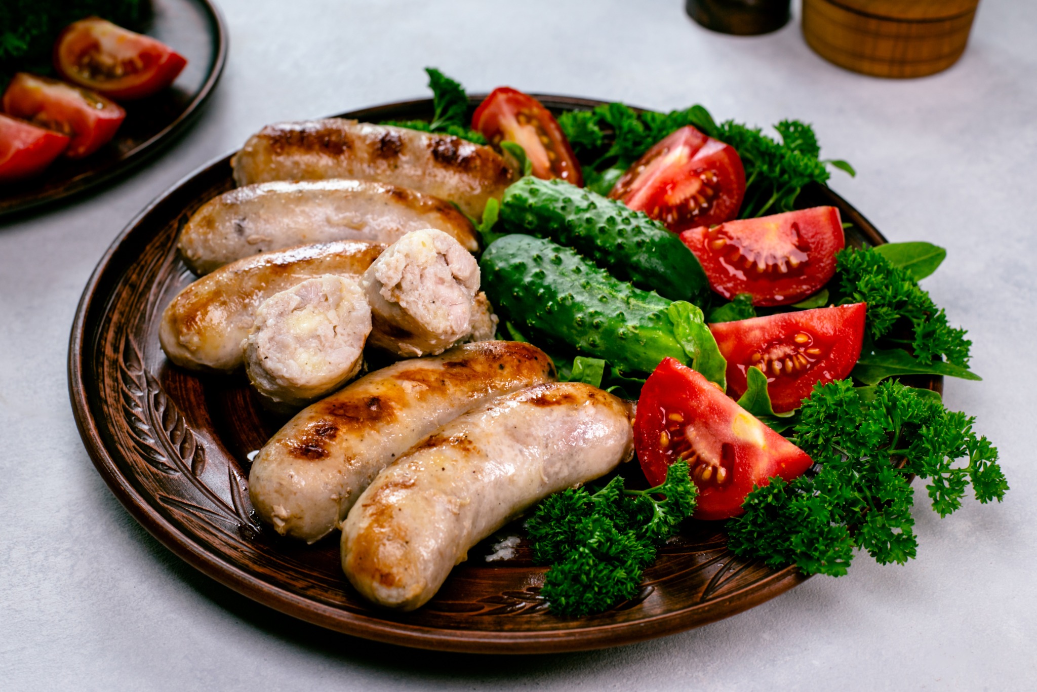 Ceramiczny talerz z grillowanymi kiełbaskami, świeżymi ogórkami, pomidorami i natką pietruszki. Kiełbaski są przekrojone, ukazując farsz. Całość w ciepłej tonacji.