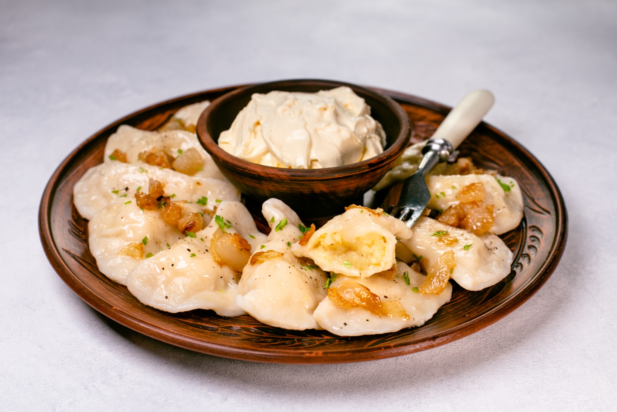 Ręcznie lepione pierogi na rustykalnym talerzu, polane skwarkami i posypane zieleniną. Obok miseczka ze śmietaną i widelec. Idealne danie na catering.