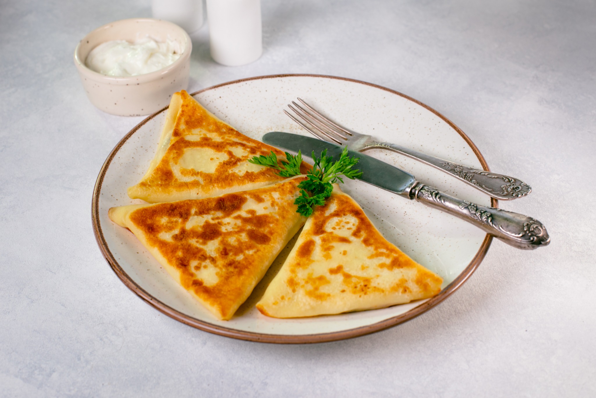 Trzy złociste naleśniki trójkątne na talerzu z rustykalnym brzegiem, udekorowane zieloną pietruszką, obok sztućce i miseczka z sosem. Jasne tło.